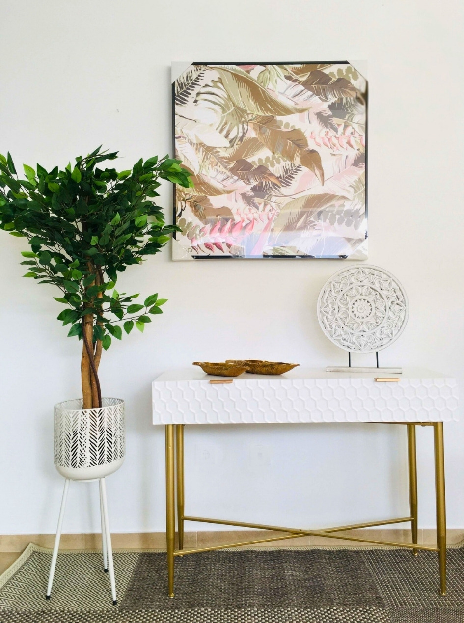 Console branco com pernas douradas, planta em vaso branco e quadro decorativo de folhas