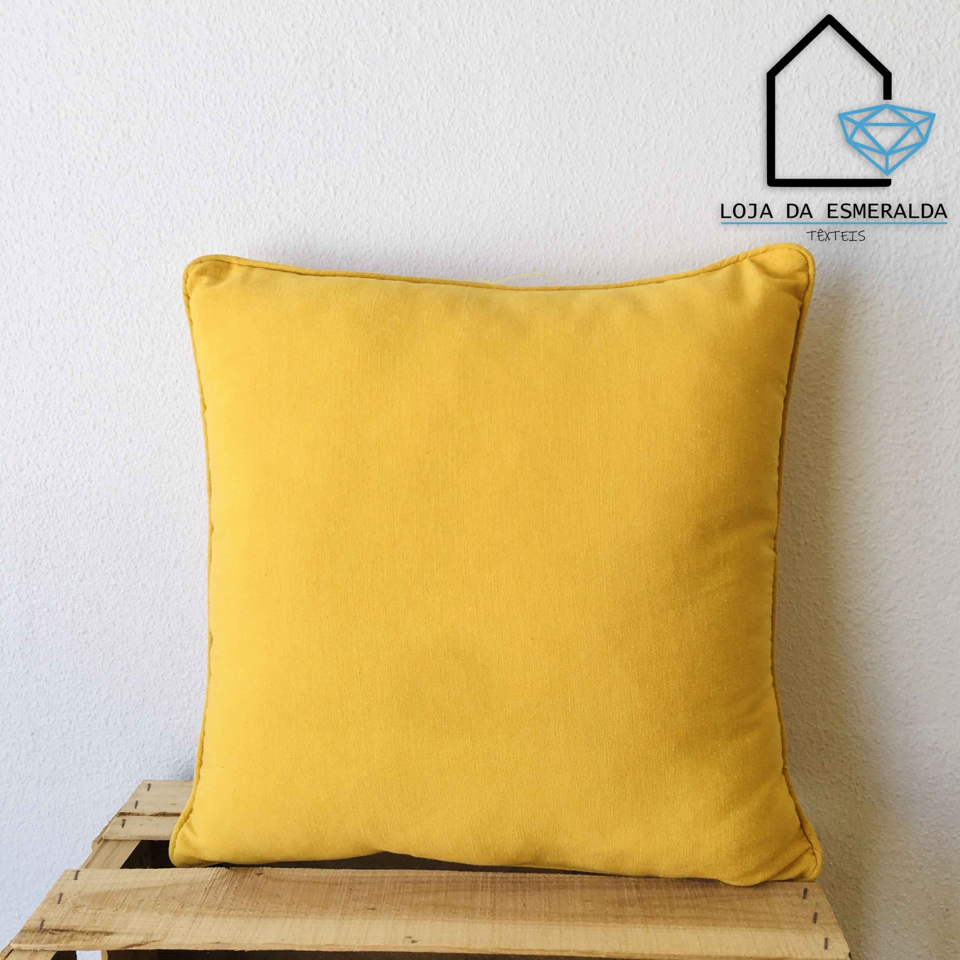 Almofada quadrada amarela sobre superfície de madeira com logótipo da Loja da Esmeralda Têxteis