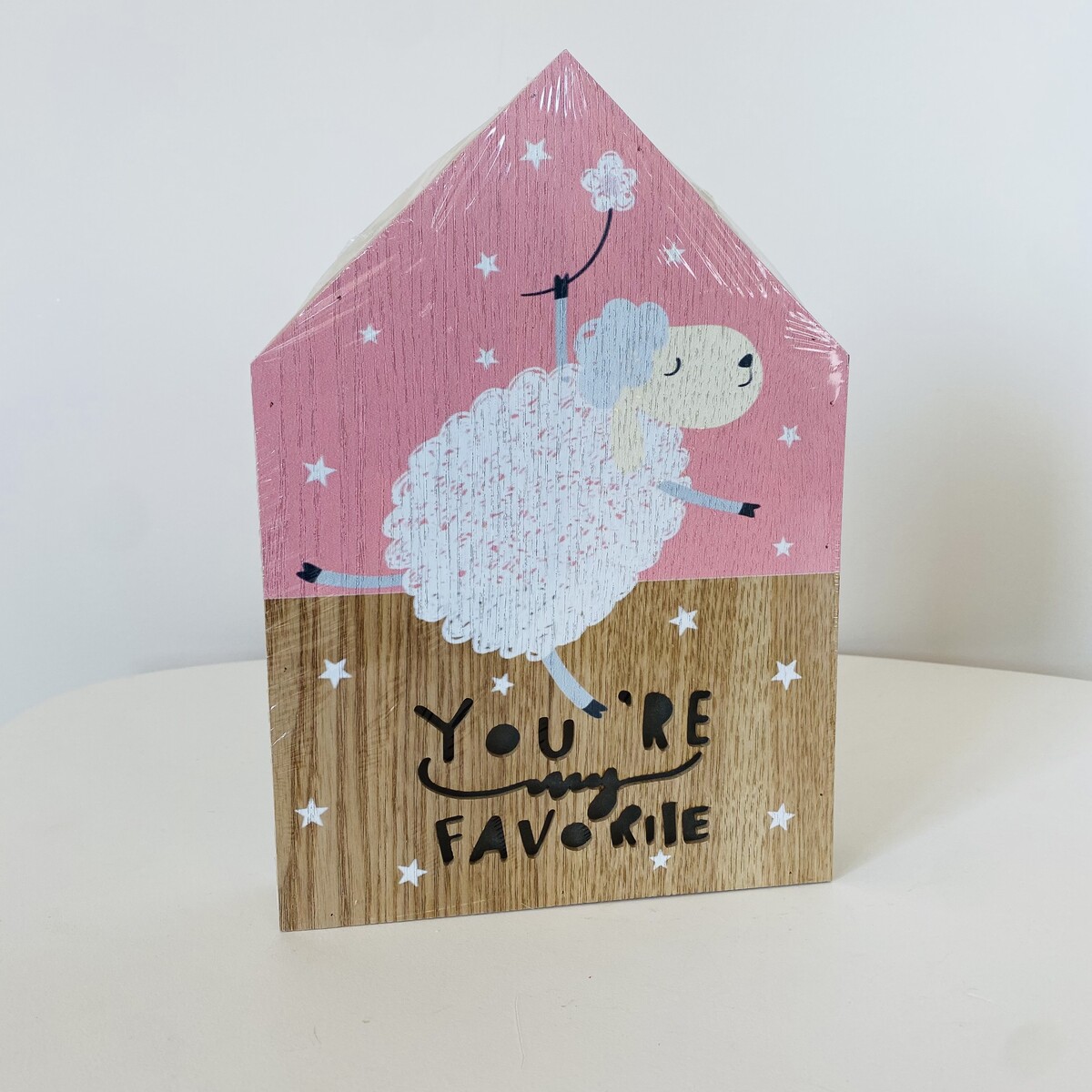 Bloco decorativo em forma de casa com ovelha e texto em fundo rosa e bege