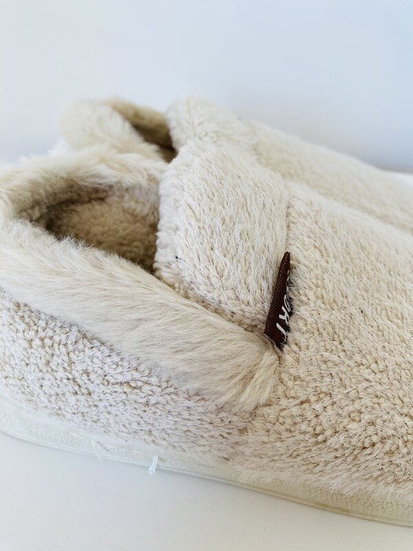 Pantufa castanha clara felpuda com etiqueta castanha e sola branca numa superfície branca