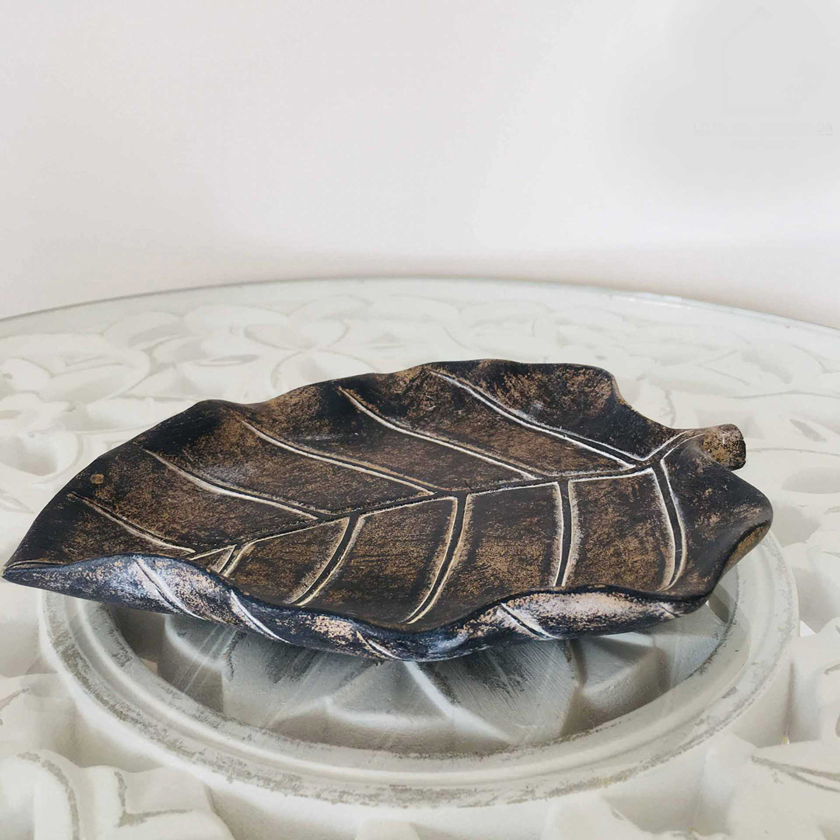 Prato decorativo em forma de folha metálico castanho e preto sobre mesa branca com relevo