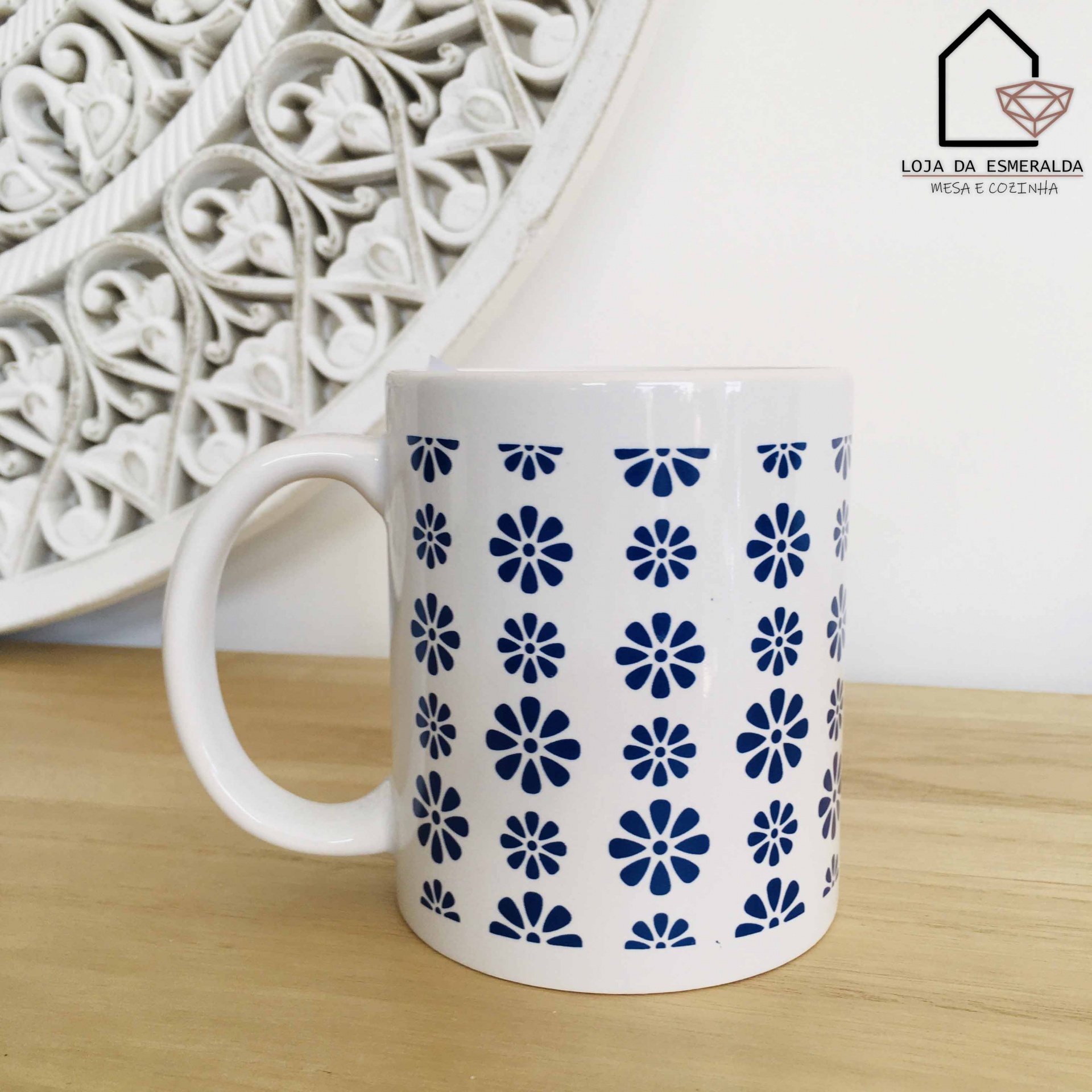 Caneca branca com padrão floral azul e logótipo de loja ao fundo