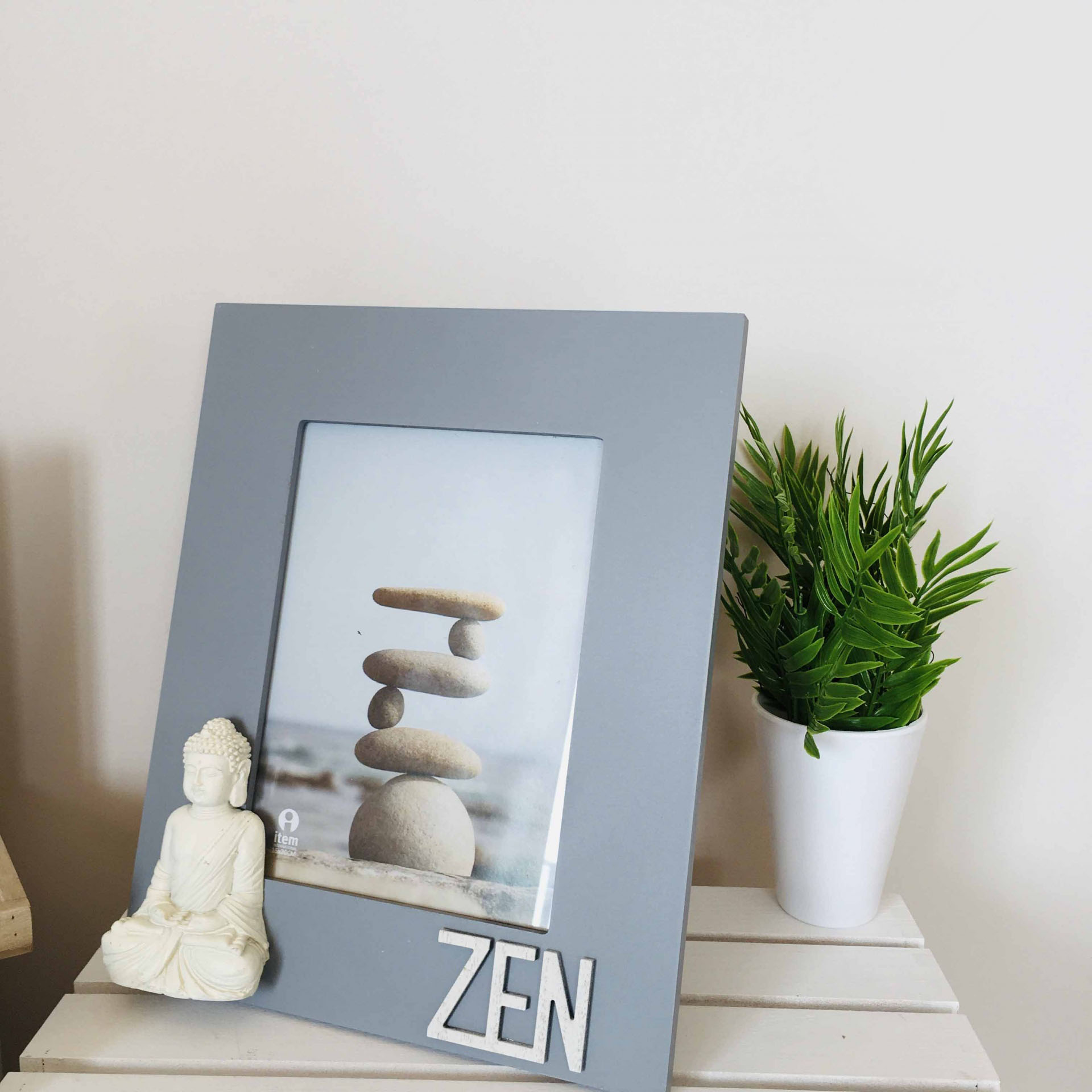 Moldura cinzenta com palavra ZEN, estátua de Buda branca e planta em vaso branco