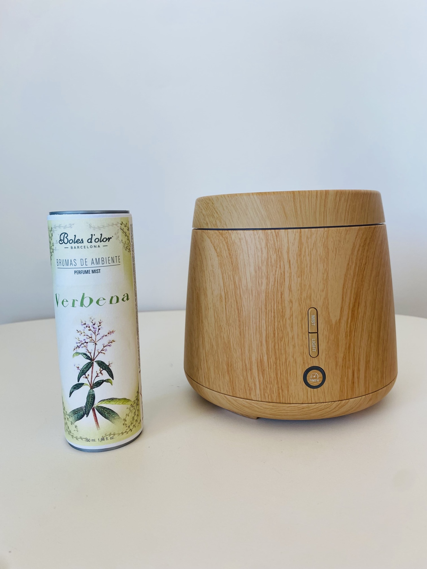 Difusor de aromas de madeira clara ao lado de frasco cilíndrico de essência Verbena sobre mesa branca