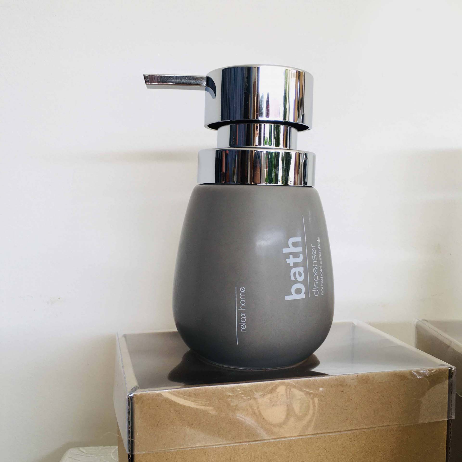 Dispenser cinzento fosco com bomba metálica prateada e texto branco, sobre caixa de papelão