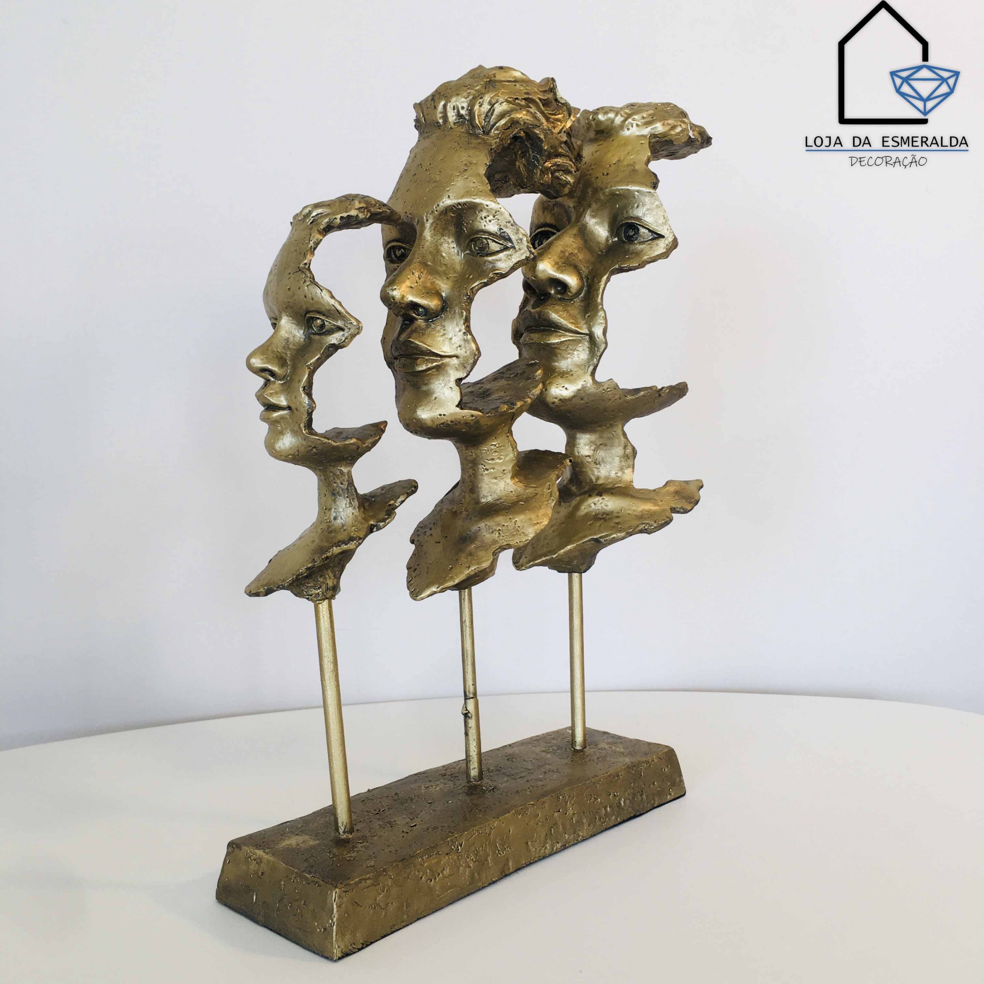 Escultura de três rostos humanos em bronze sobre base retangular com logótipo ao fundo