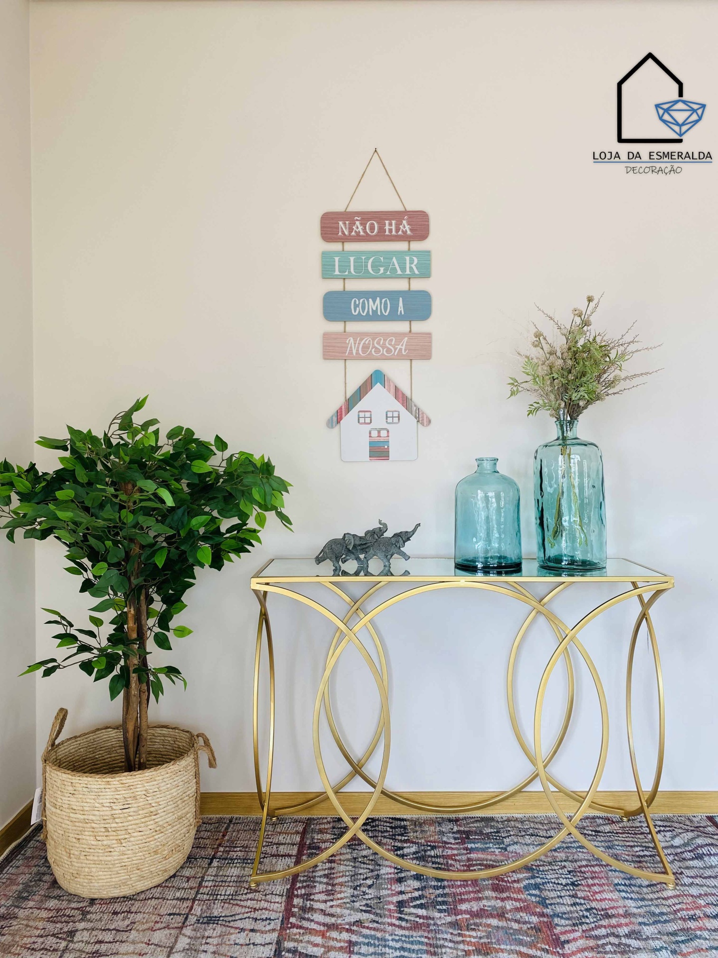 Consola decorativa dourada com tampo de vidro, vasos de vidro azul, estatueta de rinocerontes e planta artificial ao lado, placa pendurada na parede com texto em português.