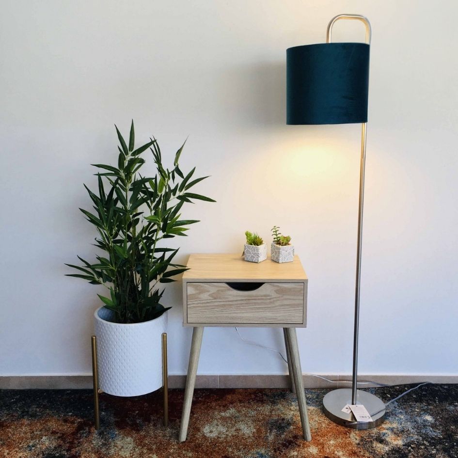 Mesa de cabeceira com gaveta em madeira clara, vaso branco com planta, candeeiro de pé com sombra azul escuro e 2 vasos pequenos com suculentas