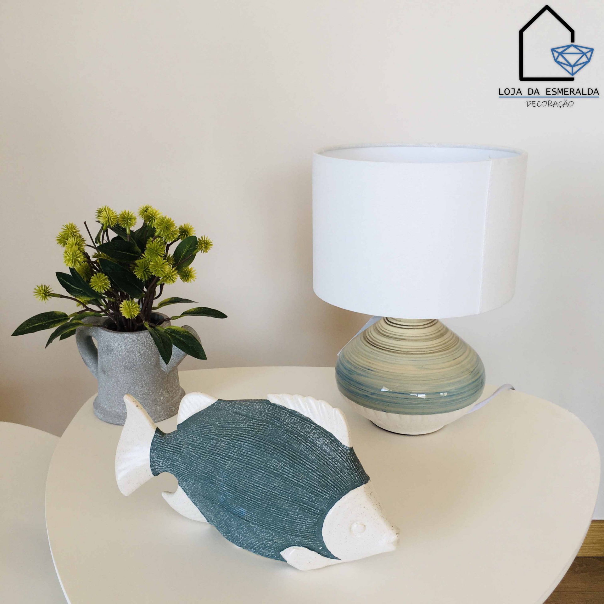 Peixe decorativo azul e branco, vaso cinza com flores e lâmpada com base azul e bege numa mesa branca