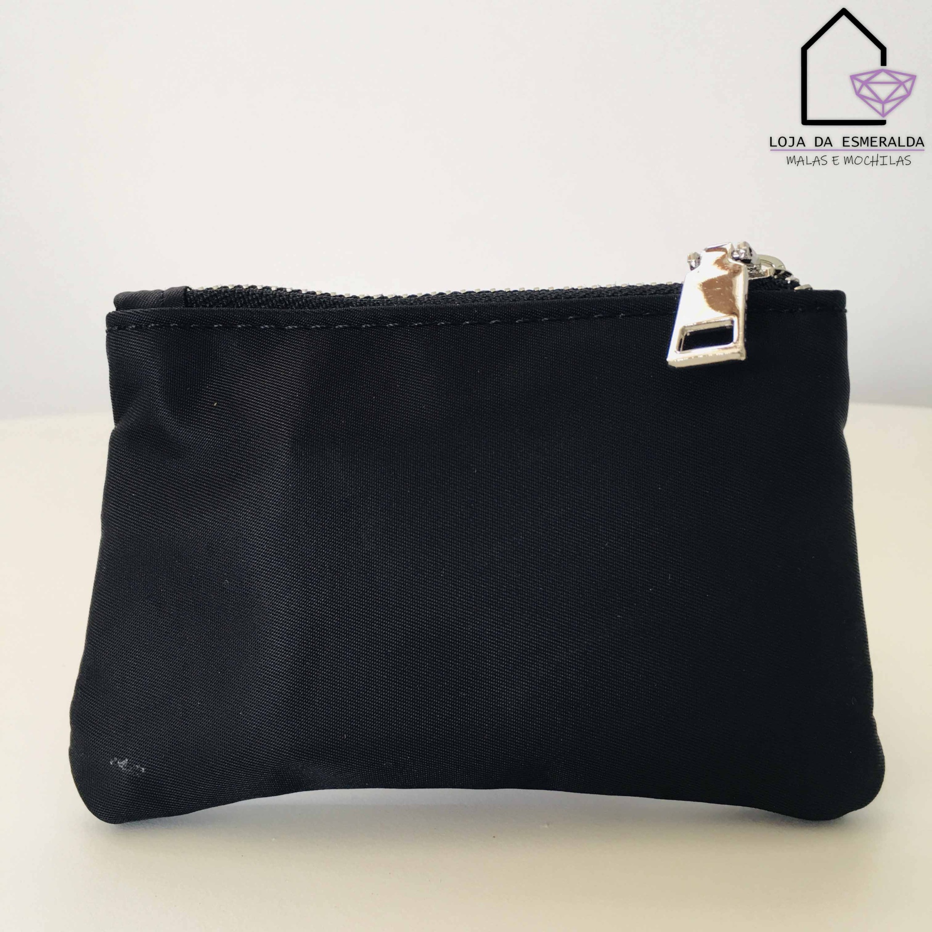 Bolsa pequena preta com fecho metálico sobre superfície clara e logótipo da loja ao fundo