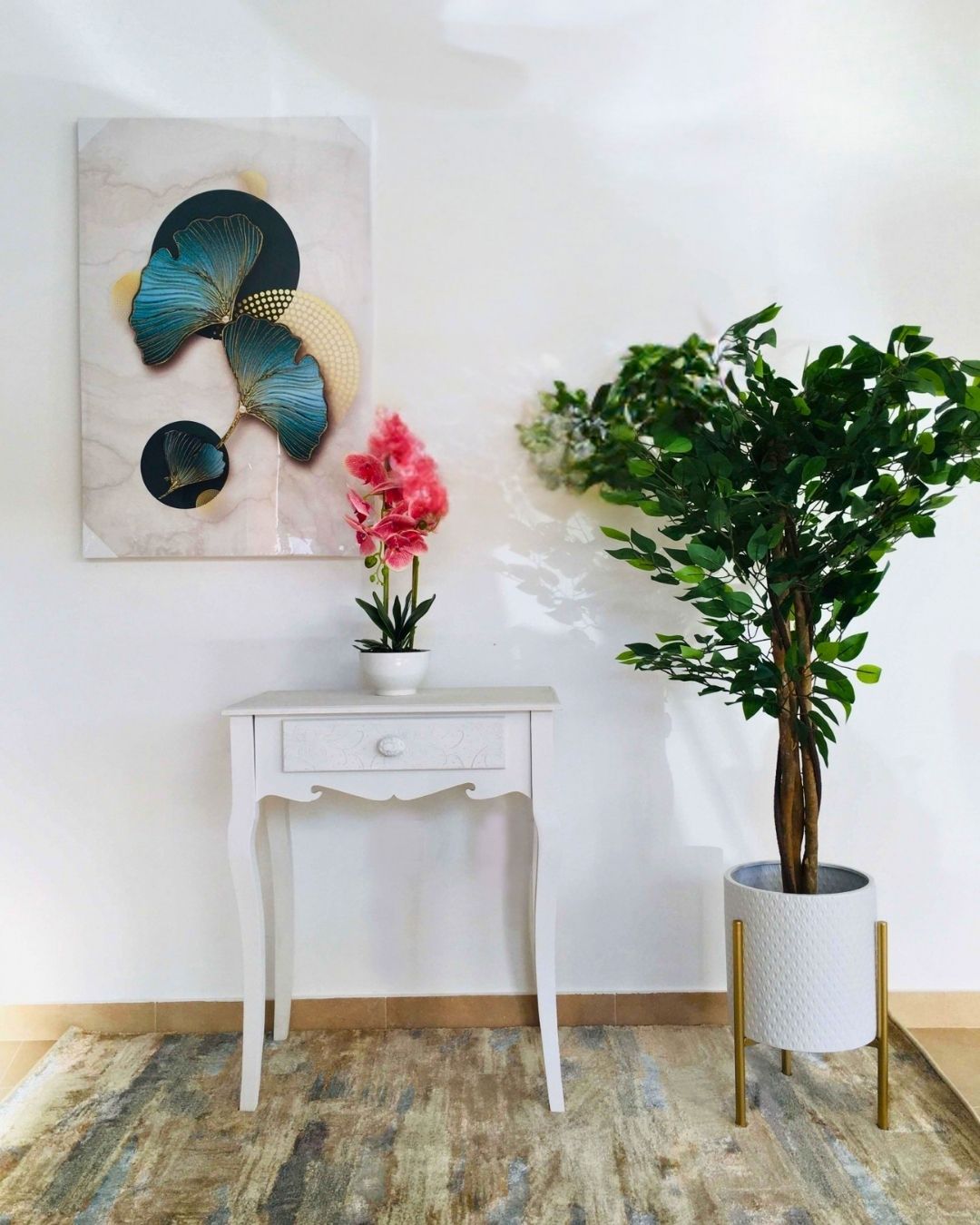 Consola branca com vaso de flores cor-de-rosa, quadro decorativo e planta verde em vaso branco com suporte dourado