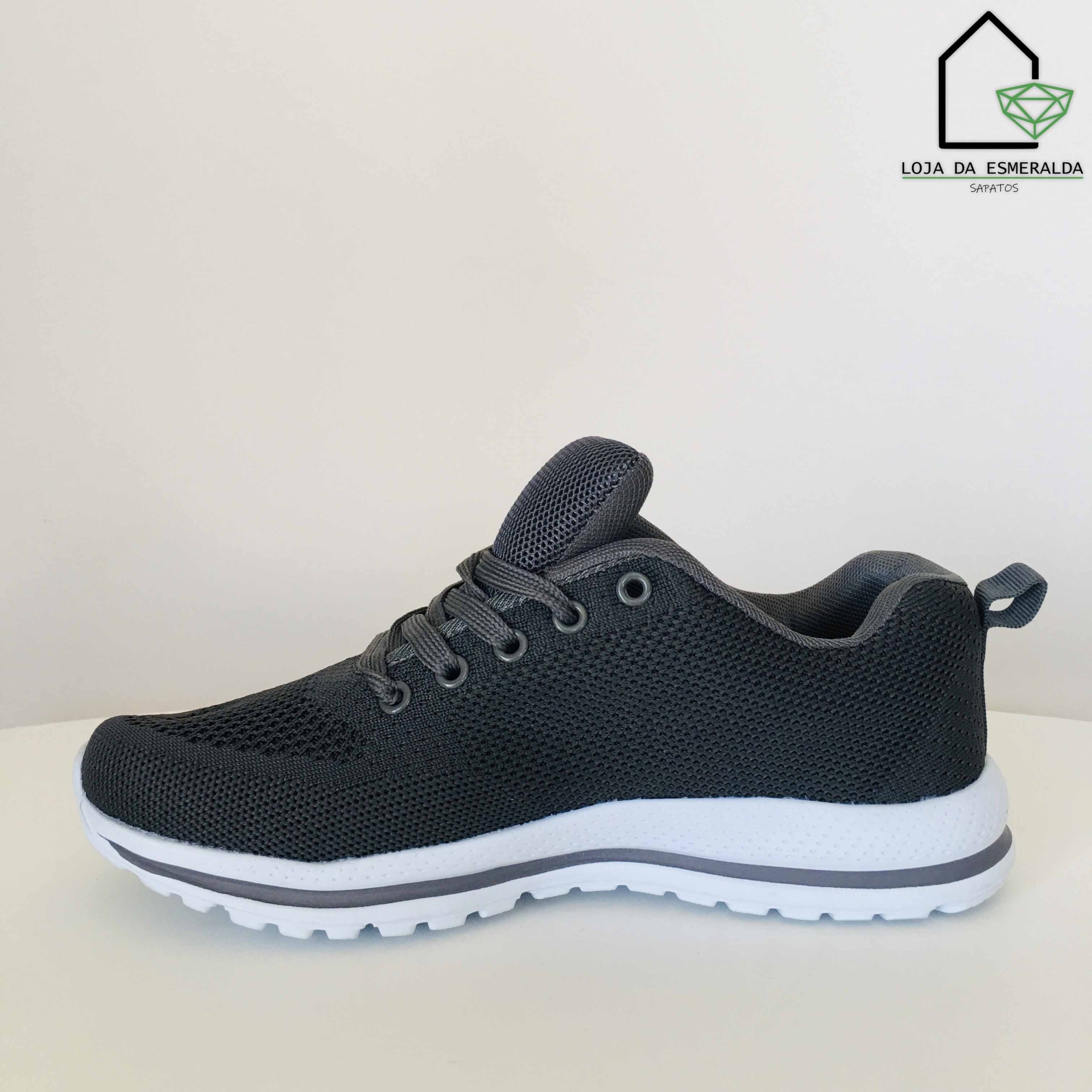 Tênis desportivo preto com sola branca e cadarços pretos em fundo branco.