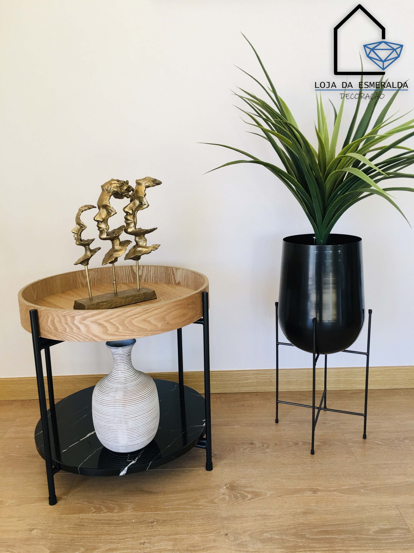 Mesa de apoio com duas prateleiras e esculturas decorativas, vaso metálico preto com planta artificial sobre chão de madeira