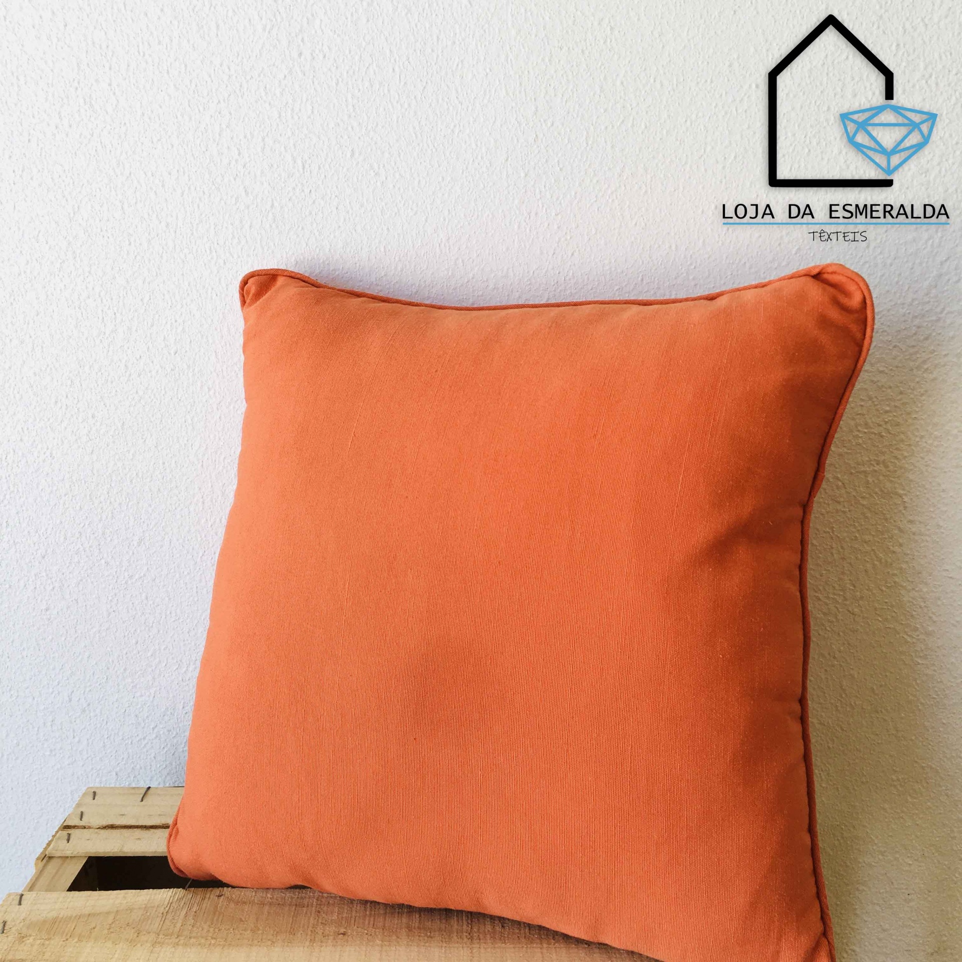 Almofada laranja quadrada sobre madeira com fundo branco e logotipo Loja da Esmeralda