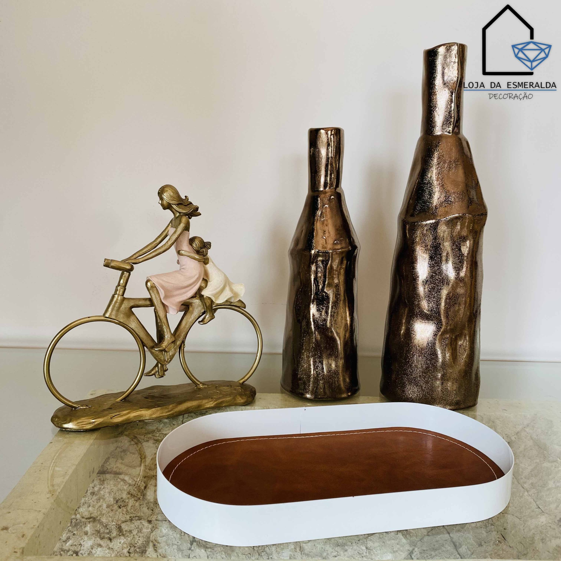 Escultura metálica de mulher e criança em velocípede, vasos bronze e tabuleiro branco e castanho sobre pedra clara.