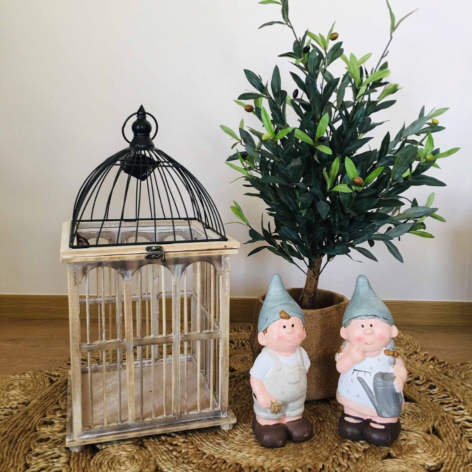 Gaiola decorativa em madeira e metal, duas figuras de gnomos e plantas artifíciais verdes em vaso castanho, num tapete de corda