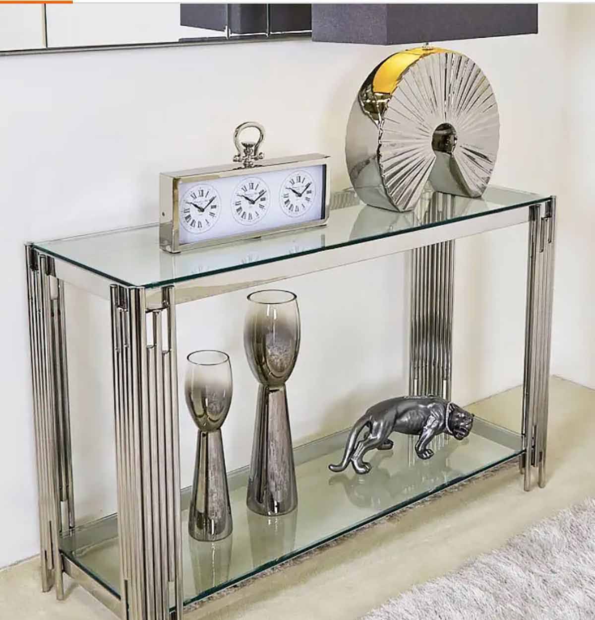Consola metálica cromada com tampo e prateleira de vidro, relógio de três fusos, taças metálicas, escultura de pantera e peça decorativa redonda.