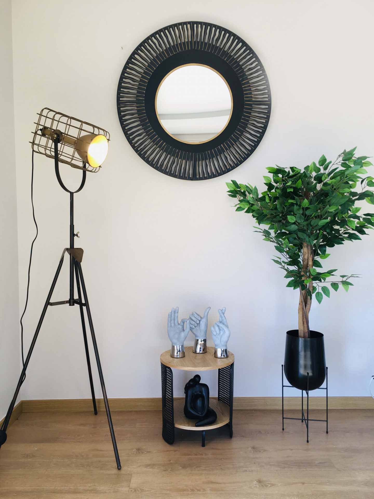 Decoração interior com candeeiro de pé, espelho redondo, mesa com esculturas de mãos e planta em floreira preta