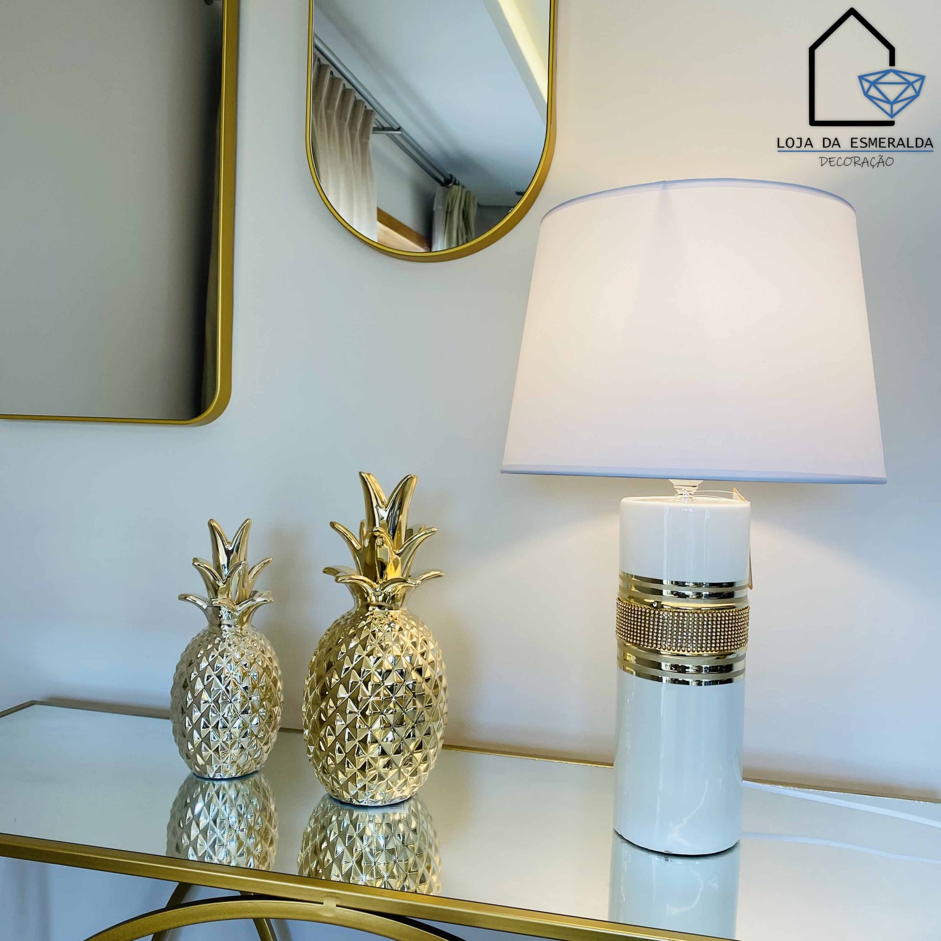 Conjunto decorativo com abacaxis dourados e candeeiro branco com detalhes dourados sobre mesa branca com moldura dourada