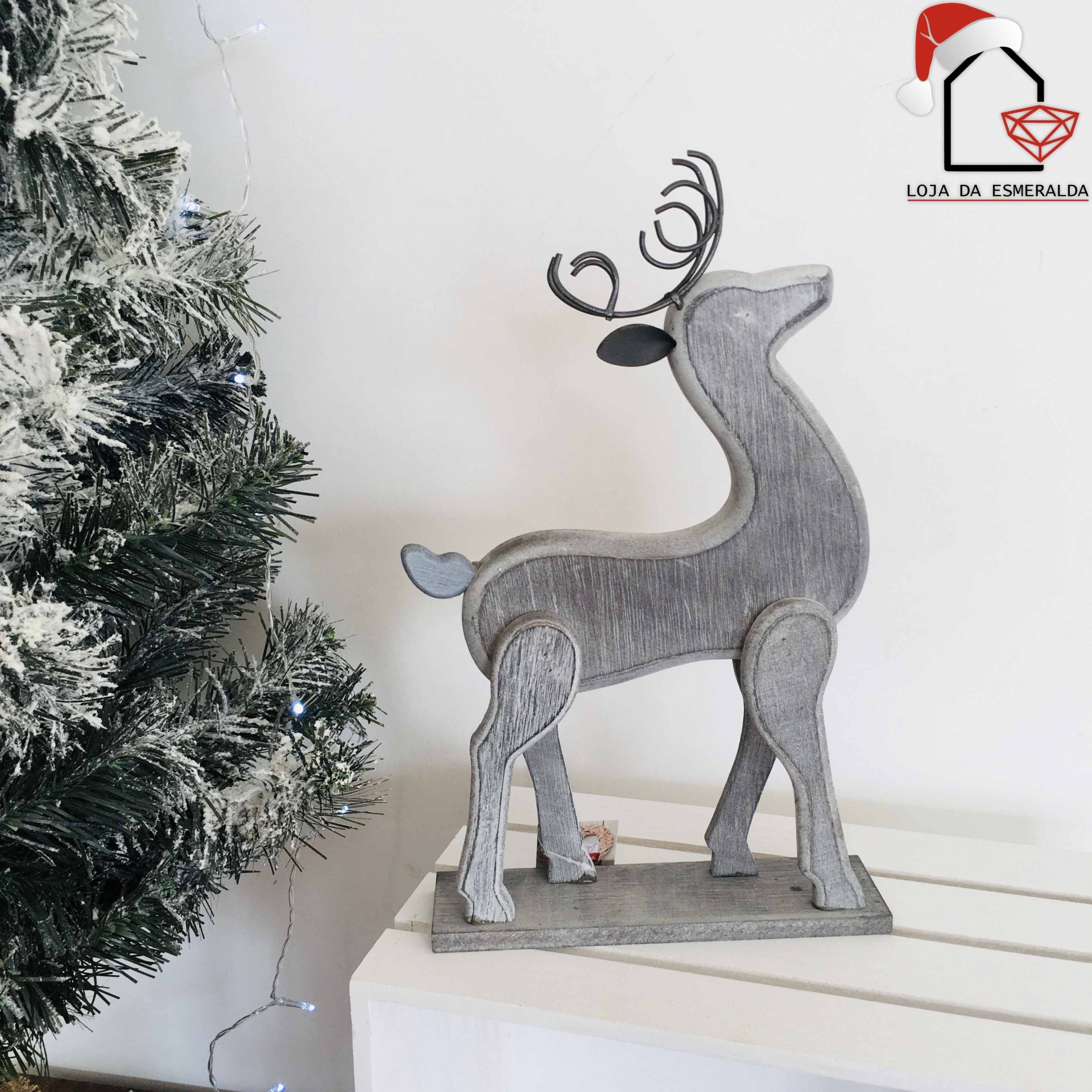 Figurinha decorativa de rena cinzenta em madeira ao lado de árvore de Natal artificial com neve