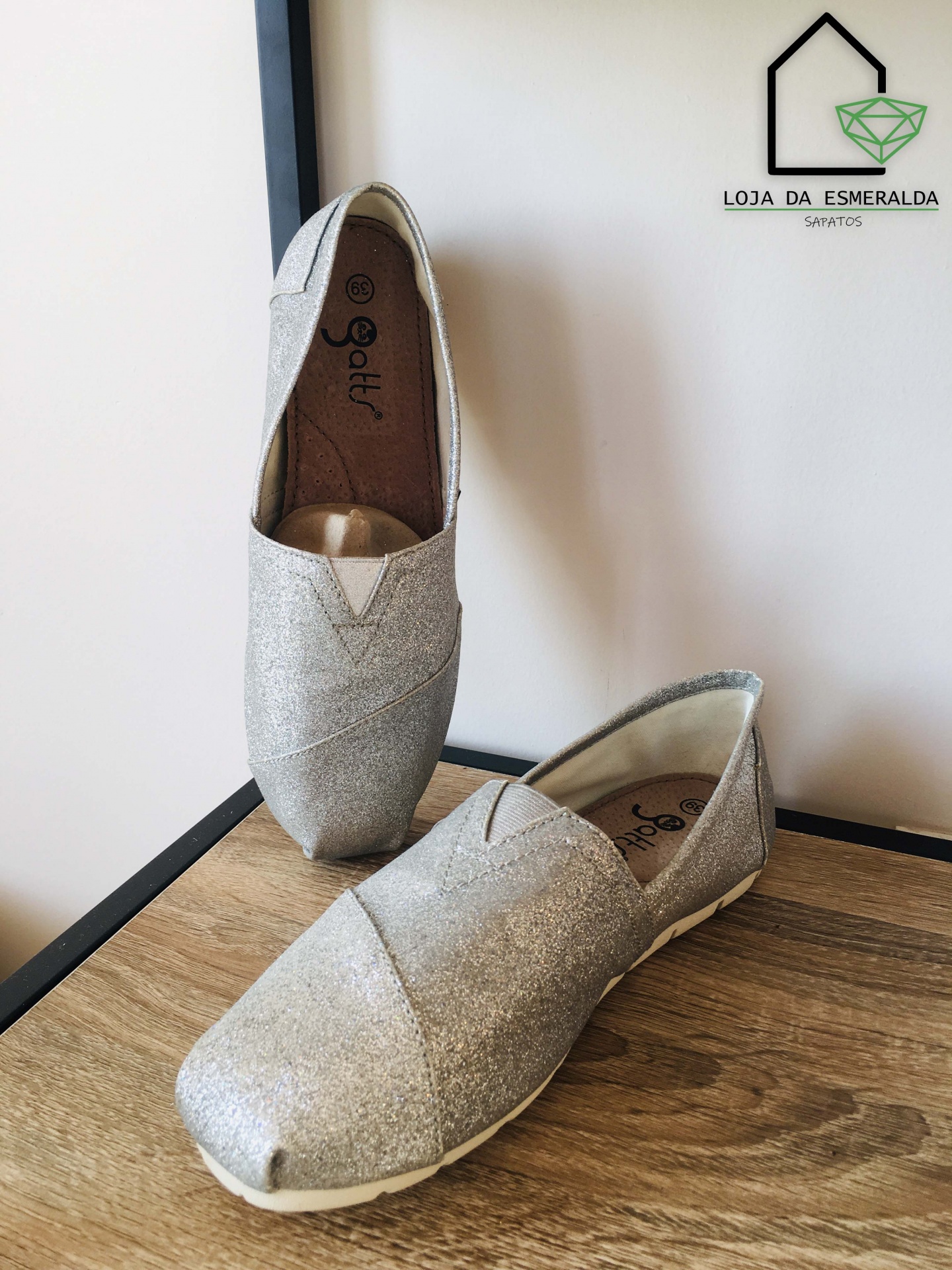 Par de sapatos prateados brilhantes tipo alpargatas na madeira ao lado do logótipo da loja