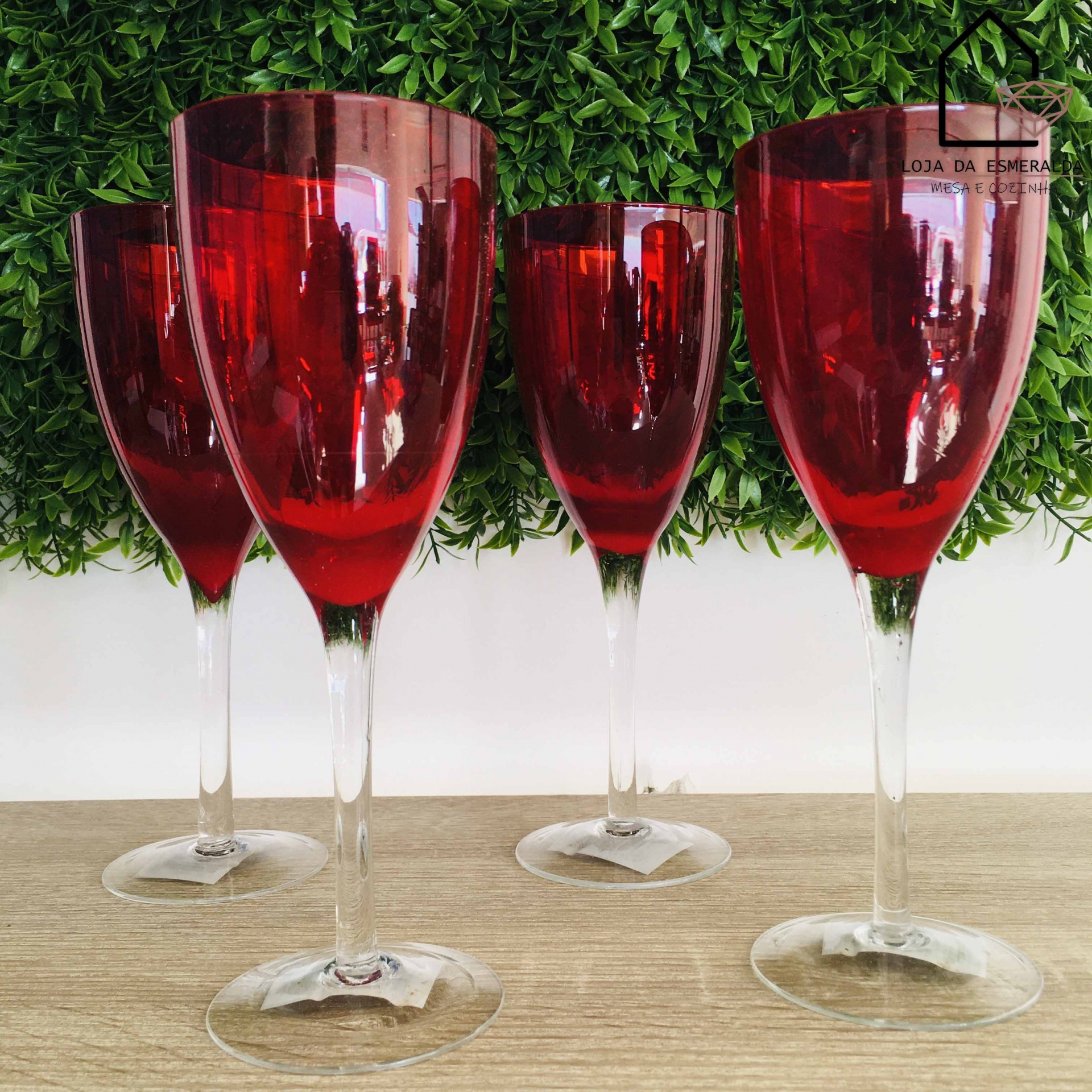 Quatro copos de vinho vermelhos com hastes claras sobre mesa de madeira clara, fundo verde de plantas artificiais