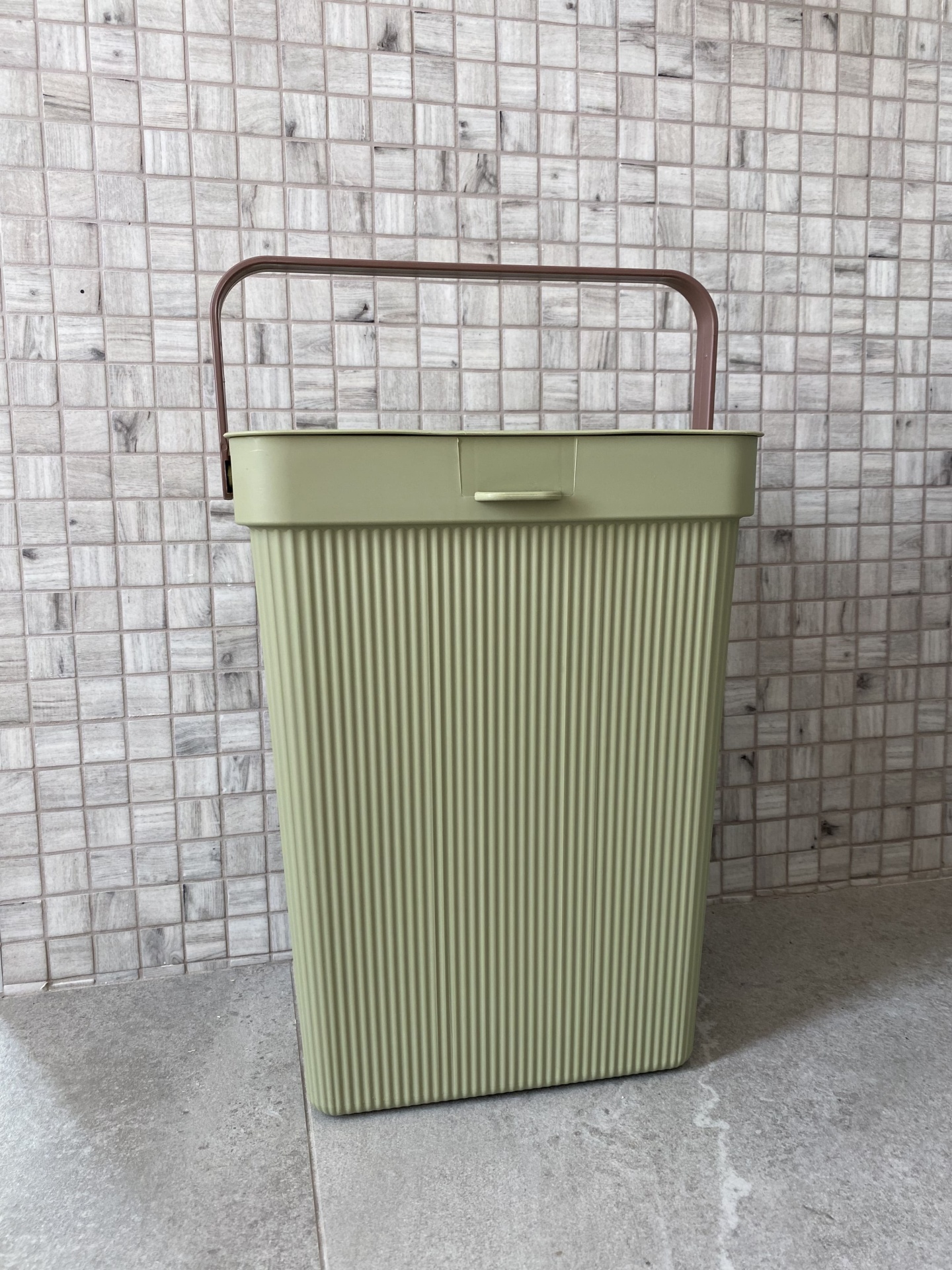 Contentor rectangular de plástico verde claro com pega castanha em chão de azulejo