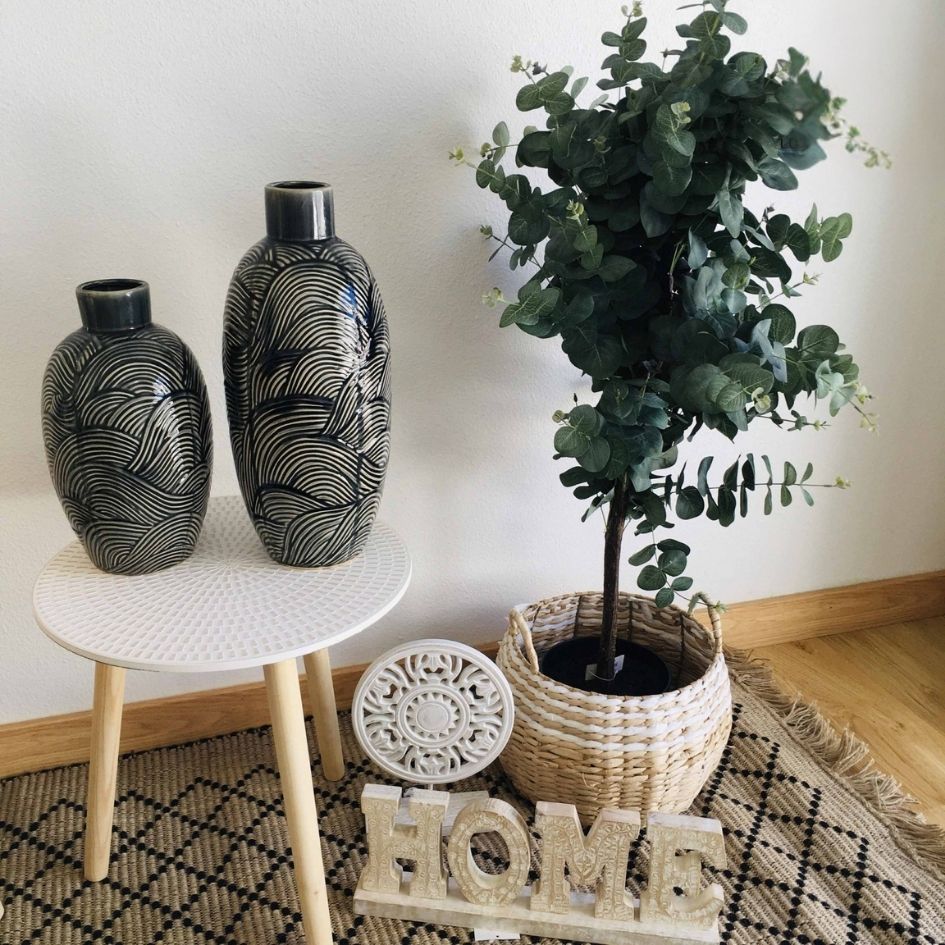 Vasos decorativos pretos com padrões brancos, planta em vaso de palha, palavra HOME em tecido, sobre tapete e perto de parede branca.