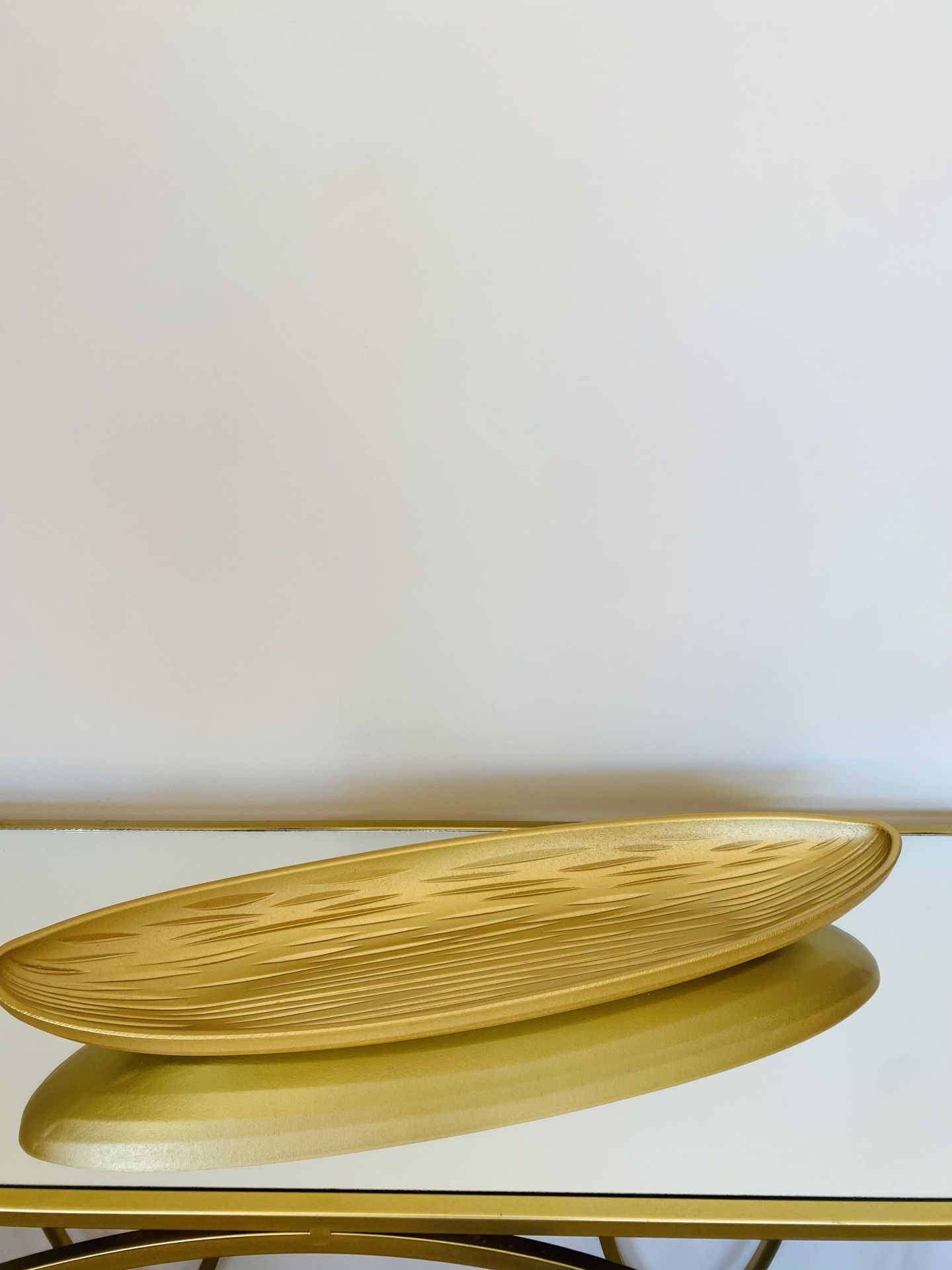 Fruteira dourada oval com relevos geométricos sobre mesa com tampo espelhado e estrutura dourada