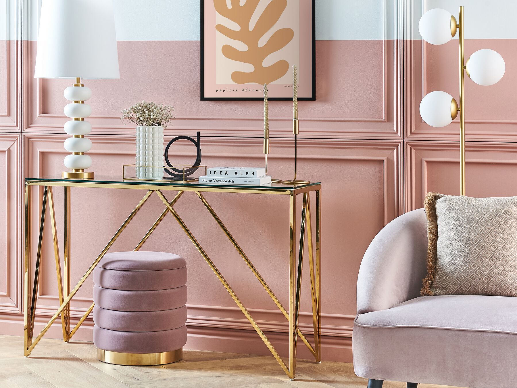 Mobiliário com candeeiro de mesa branco, mesa de vidro e estrutura dourada, banco roxo, candeeiro de pé dourado, sofá cinzento claro, parede rosa com molduras e quadro abstrato