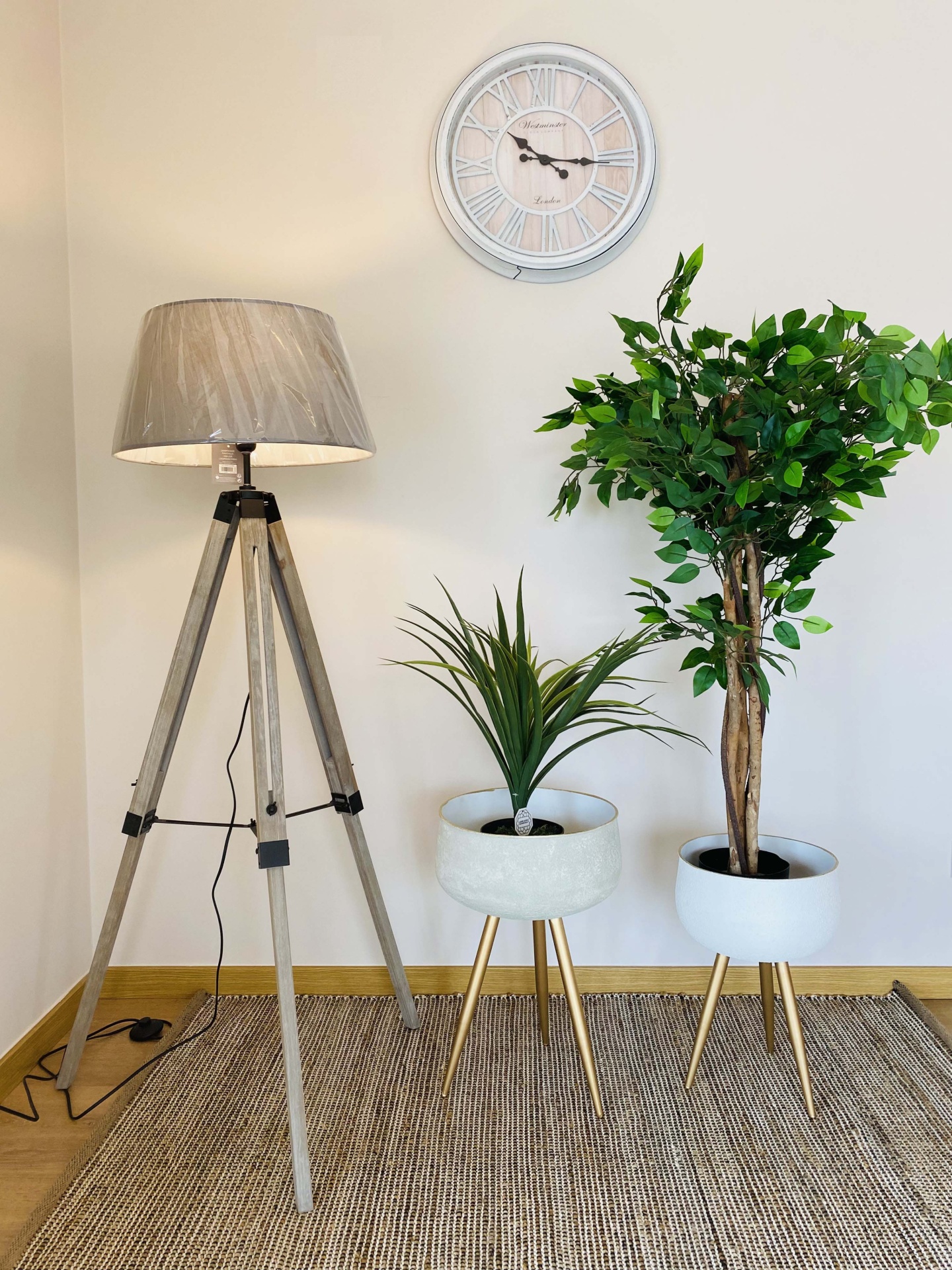 Relógio de parede branco e cinzento, lâmpada de pé com tripé de madeira e duas plantas artificiais em vasos brancos com tripés dourados