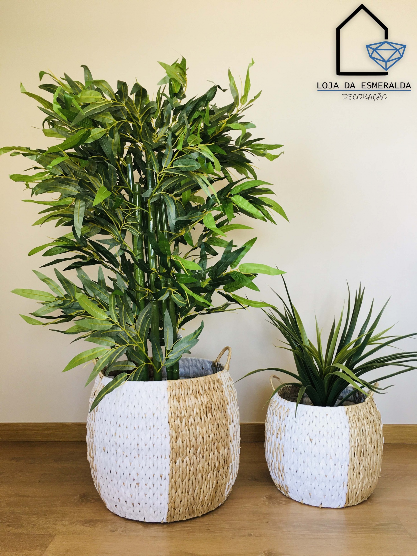 Dois vasos trançados branco e bege com plantas artificiais verdes em piso de madeira e parede clara