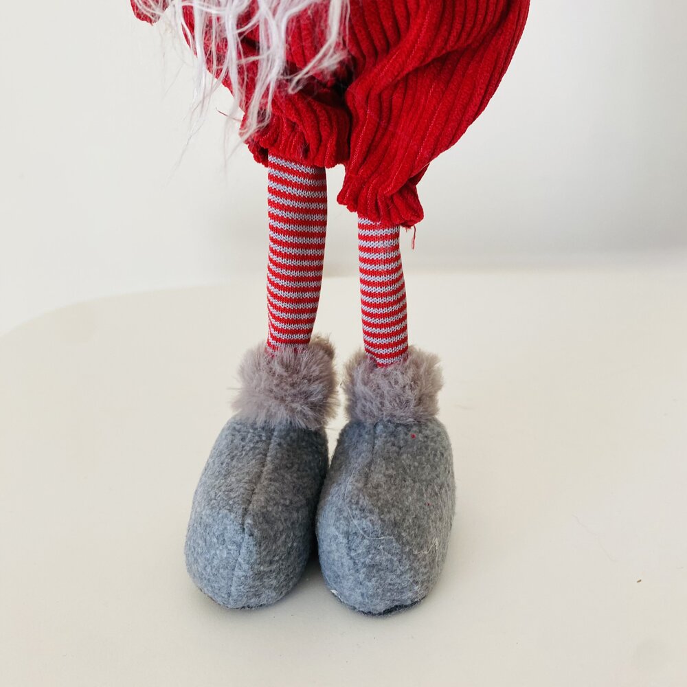 Pés de boneca com meias às riscas vermelhas e brancas, chinelos cinzentos com pelúcia e calças vermelhas.