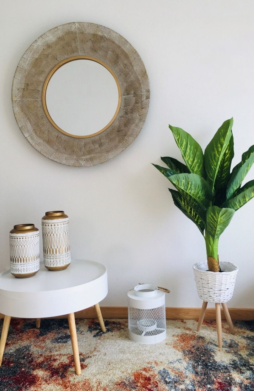 Mesa redonda branca com dois recipientes decorativos, cesto com planta e espelho redondo com moldura texturizada.