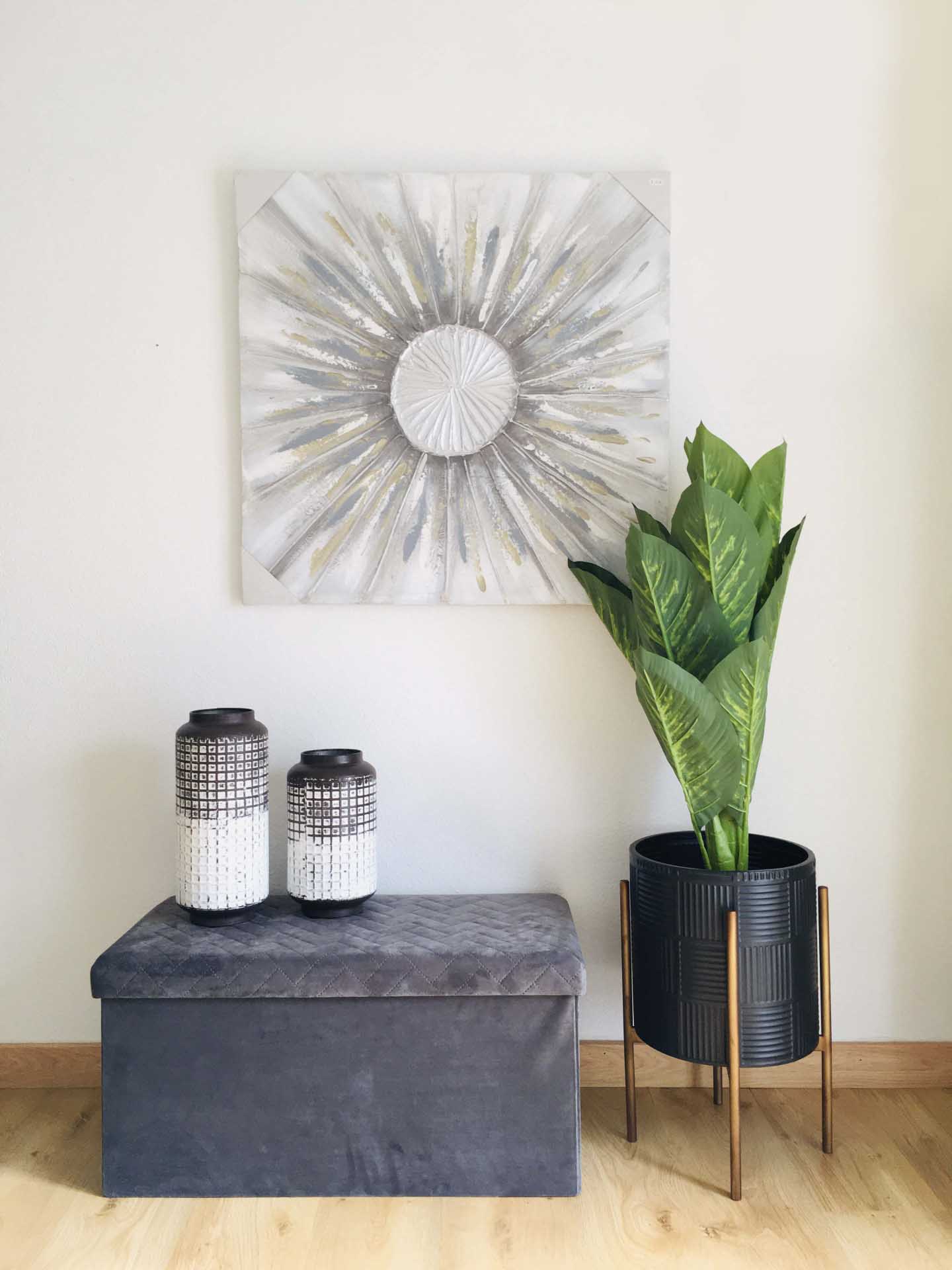 Baú cinzento com jarras decorativas e planta em vaso preto com suporte, em frente a parede com tela abstrata