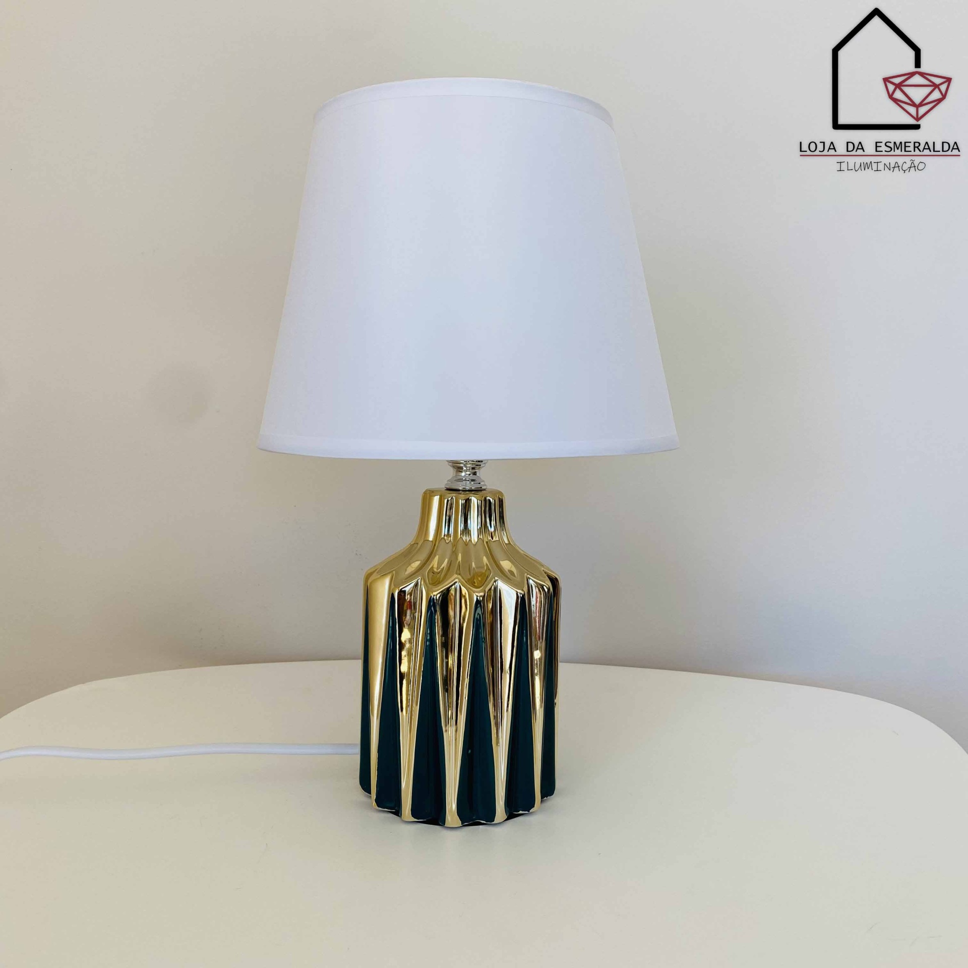 Abajur com base dourada facetada e cúpula branca sobre mesa branca