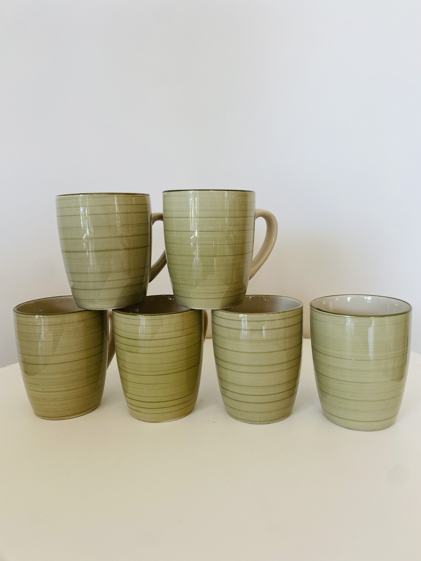 Seis chávenas cerâmicas verdes com riscas horizontais finas, duas com asa, dispostas sobre superfície branca
