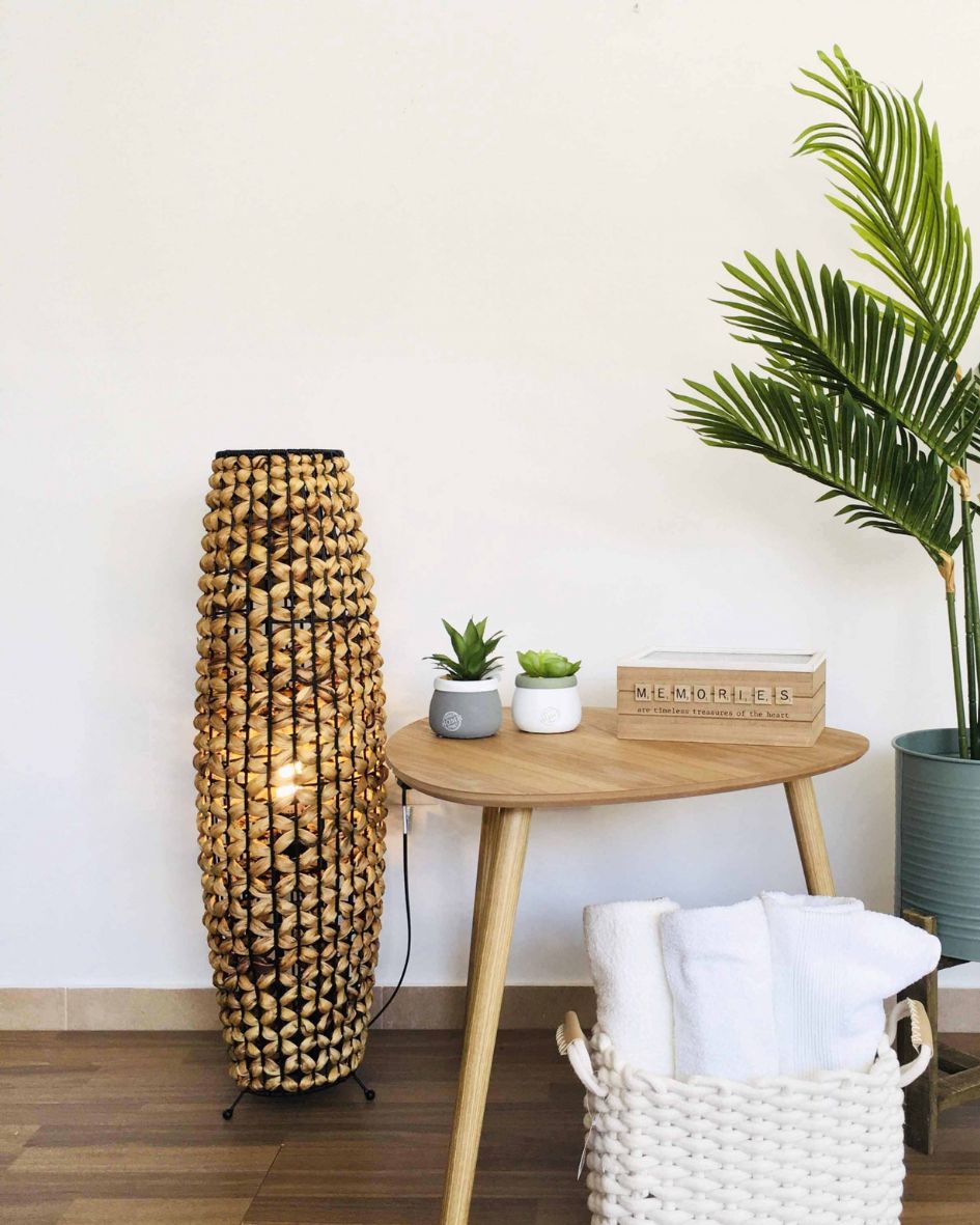 Lâmpada trançada, mesa de madeira com vasos e caixa decorativa, cesta com toalhas e planta em vaso