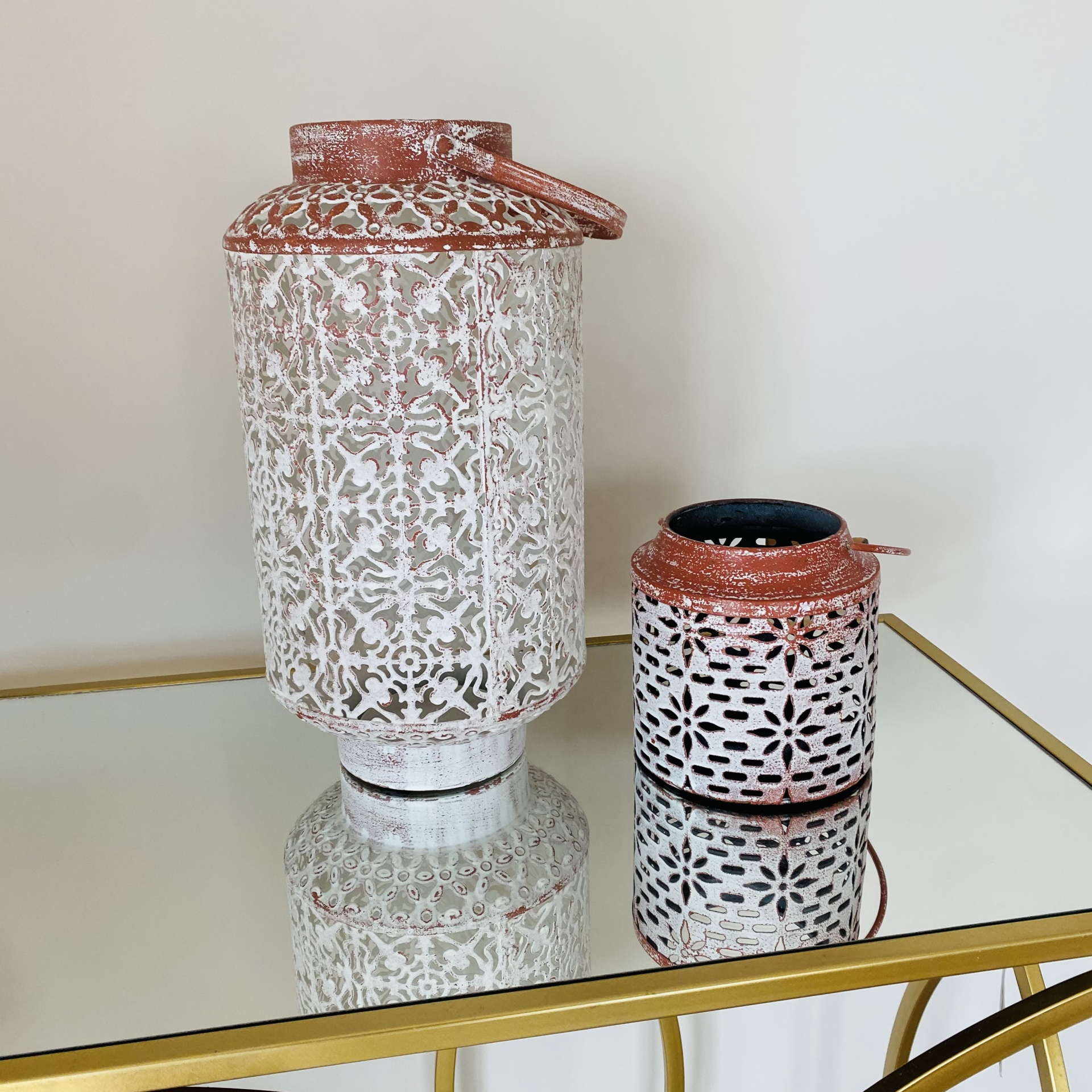 Dois lanternas decorativas em metal branco e ferrugem sobre mesa de vidro espelhado com estrutura dourada.