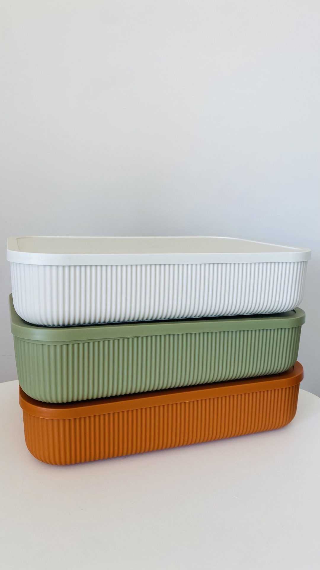 Três caixas retangulares de plástico empilhadas em branco, verde claro e castanho com textura listrada.