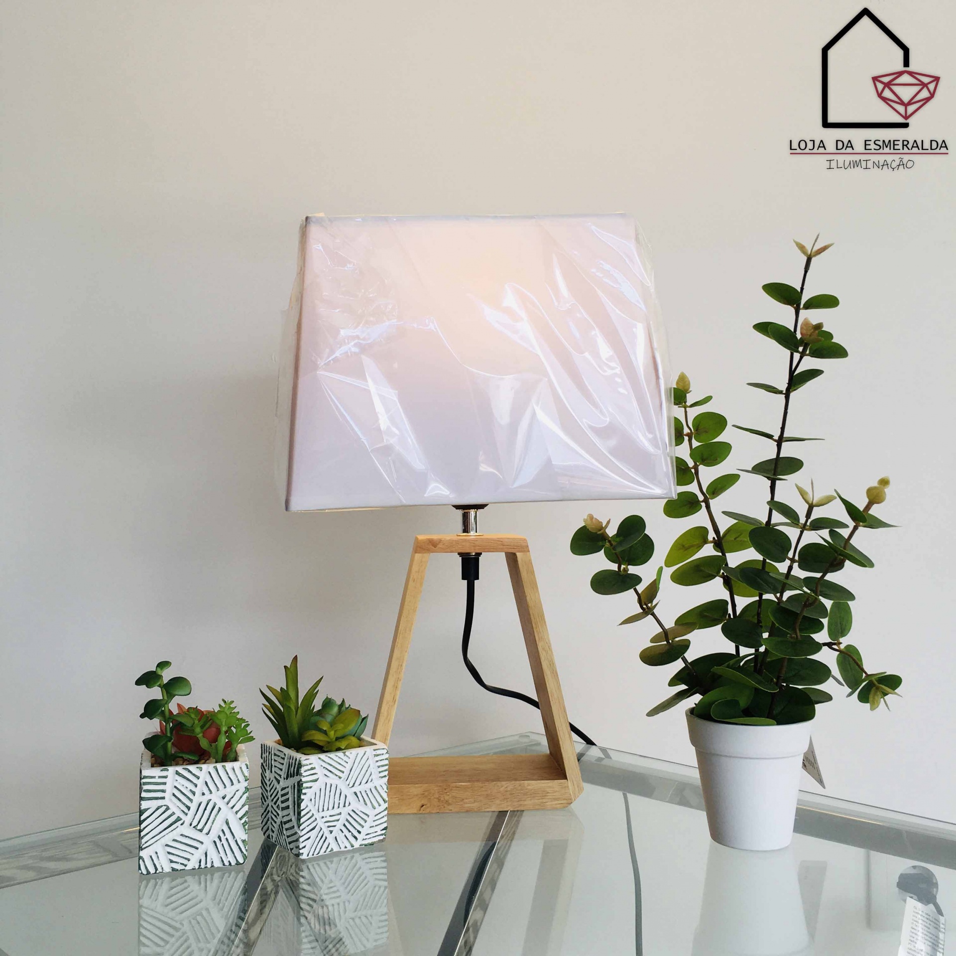 Abajur de madeira com cúpula branca em mesa de vidro com três vasos de plantas verdes