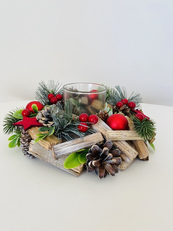 Centro de mesa natalício com paus de madeira, pinhas, ramos verdes, bagas e enfeites vermelhos com suporte de vidro no centro.