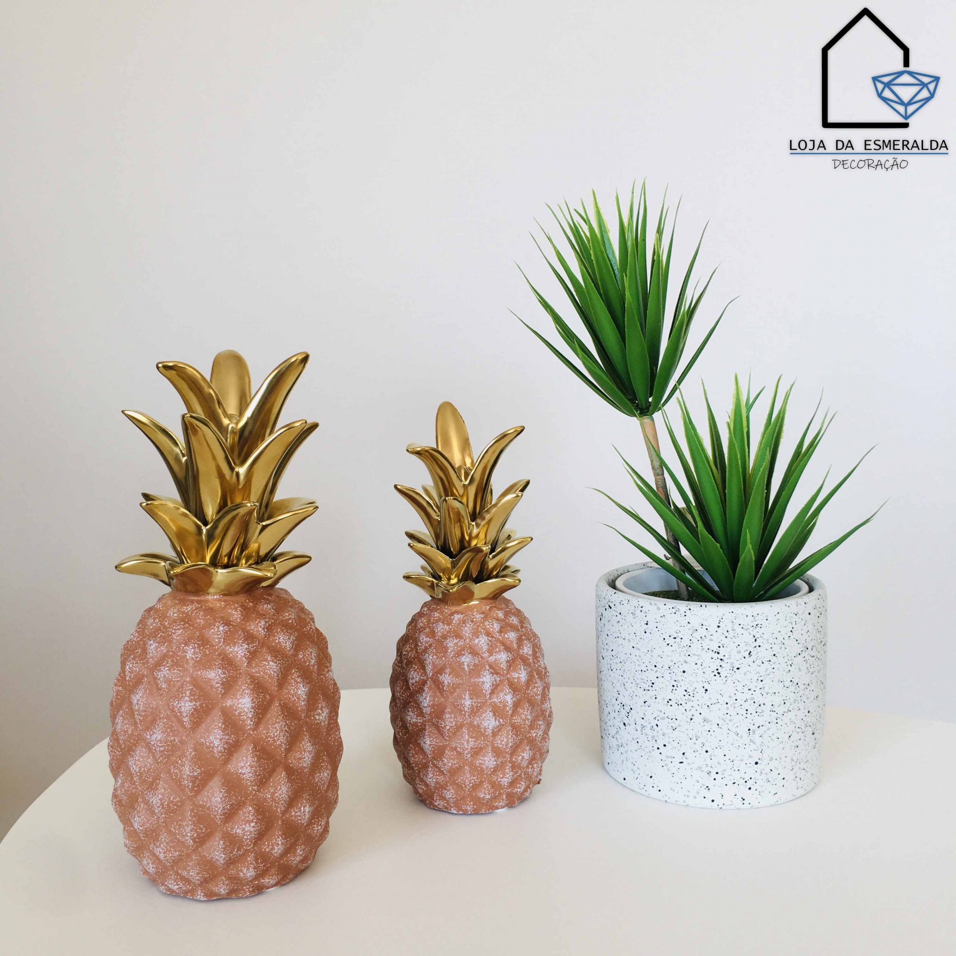 Abacaxis decorativos rosa com folhas douradas ao lado de vaso cinza com planta artificial