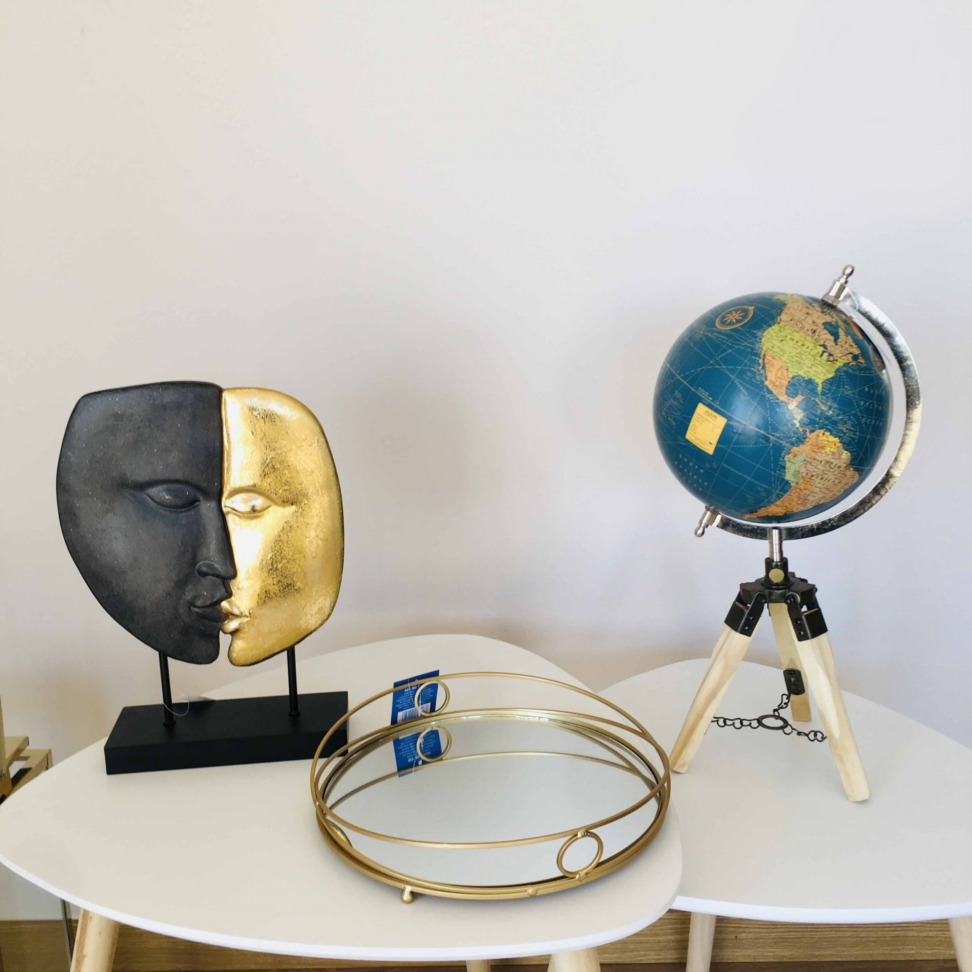 Escultura de duas faces em preto e dourado, globo azul com suporte de madeira, e espelho redondo dourado numa mesa branca