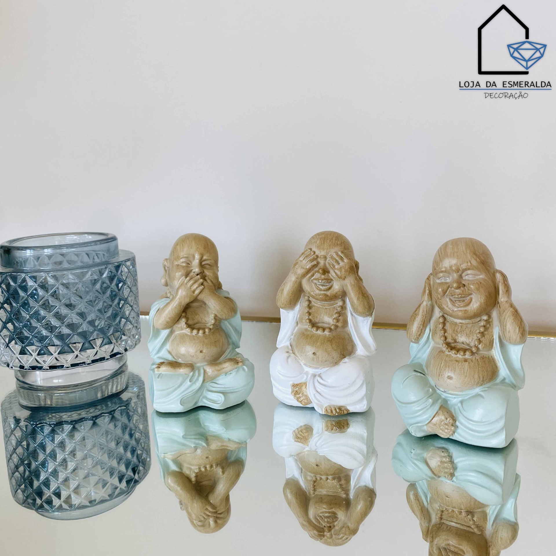 Três estátuas do Buda risonho em azul e bege junto a um castiçal cinzento numa superfície espelhada