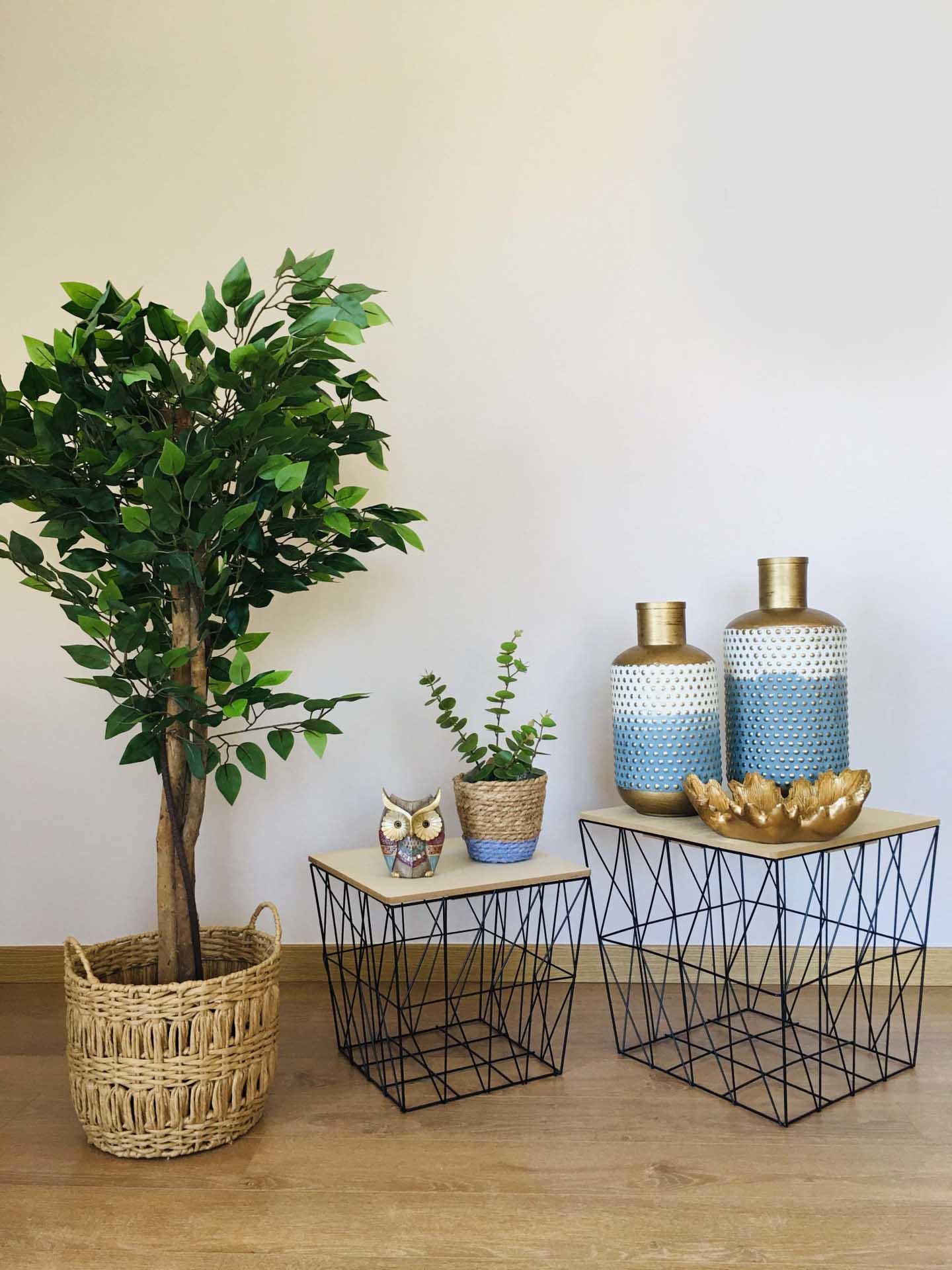 Mesas com decoração incluindo planta, garrafas cerâmicas, vaso dourado e coruja em ambiente interior.