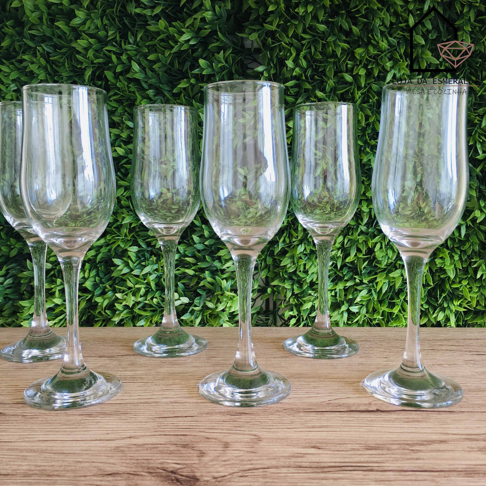 Seis copos de vidro transparentes em formatos variados sobre mesa de madeira com fundo de folhas verdes