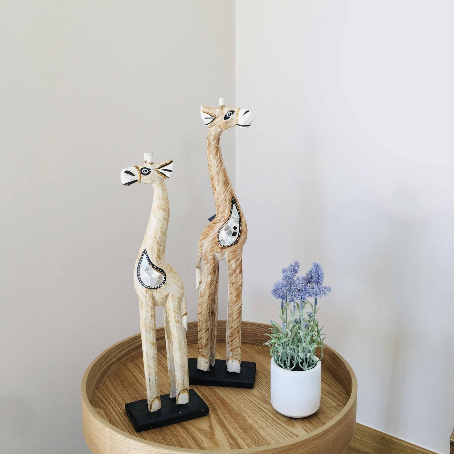 Duas esculturas de girafas em madeira clara com detalhes pretos numa mesa redonda de madeira com vaso branco e flores lilases