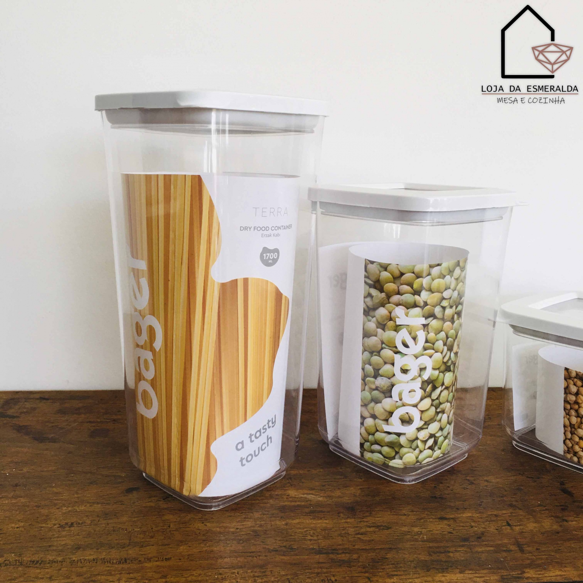 Recipientes transparentes com tampa branca contendo massas e feijão, sobre madeira.