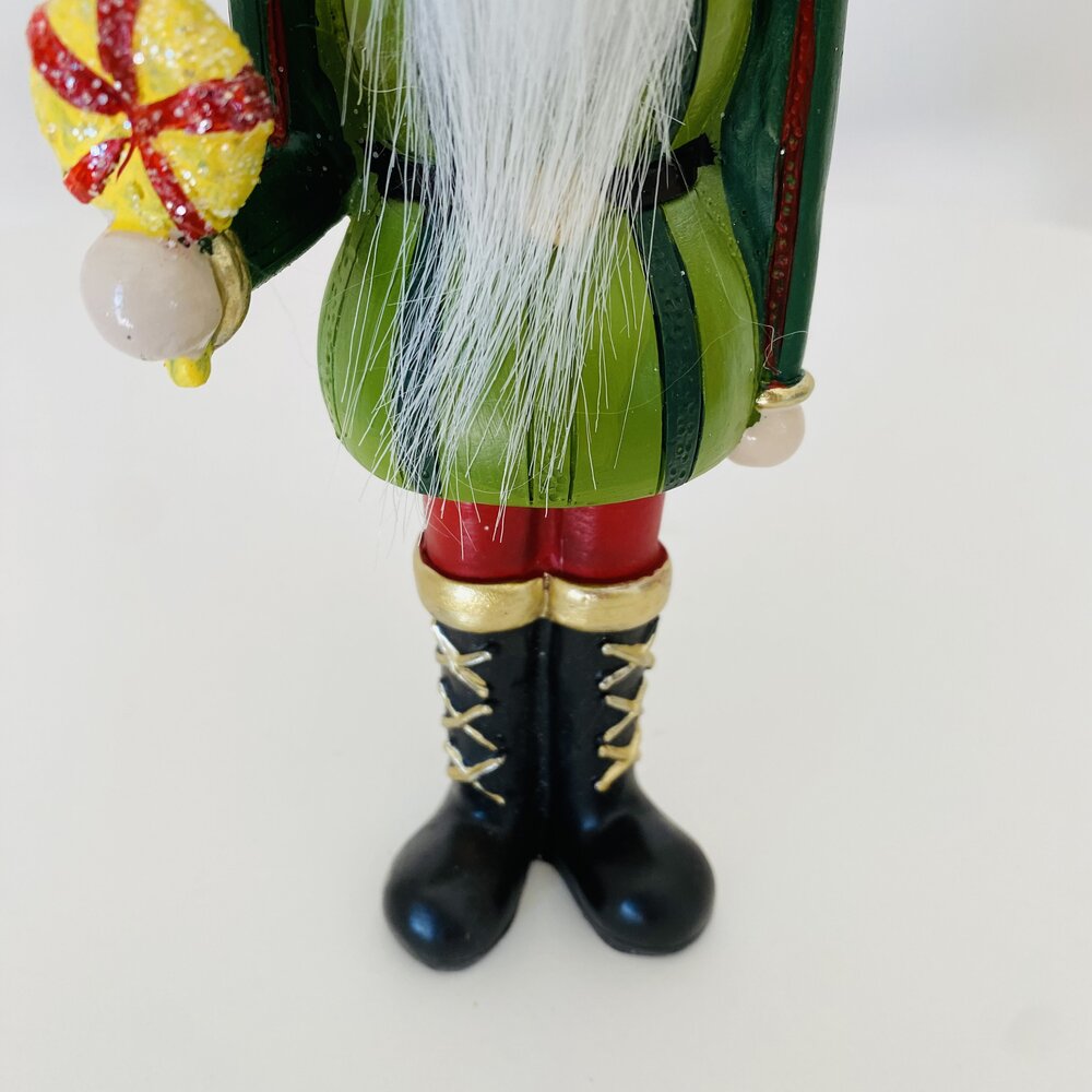 Figura decorativa de Natal com casaco verde, botas pretas com dourado, meias vermelhas, e bola decorativa amarela e vermelha